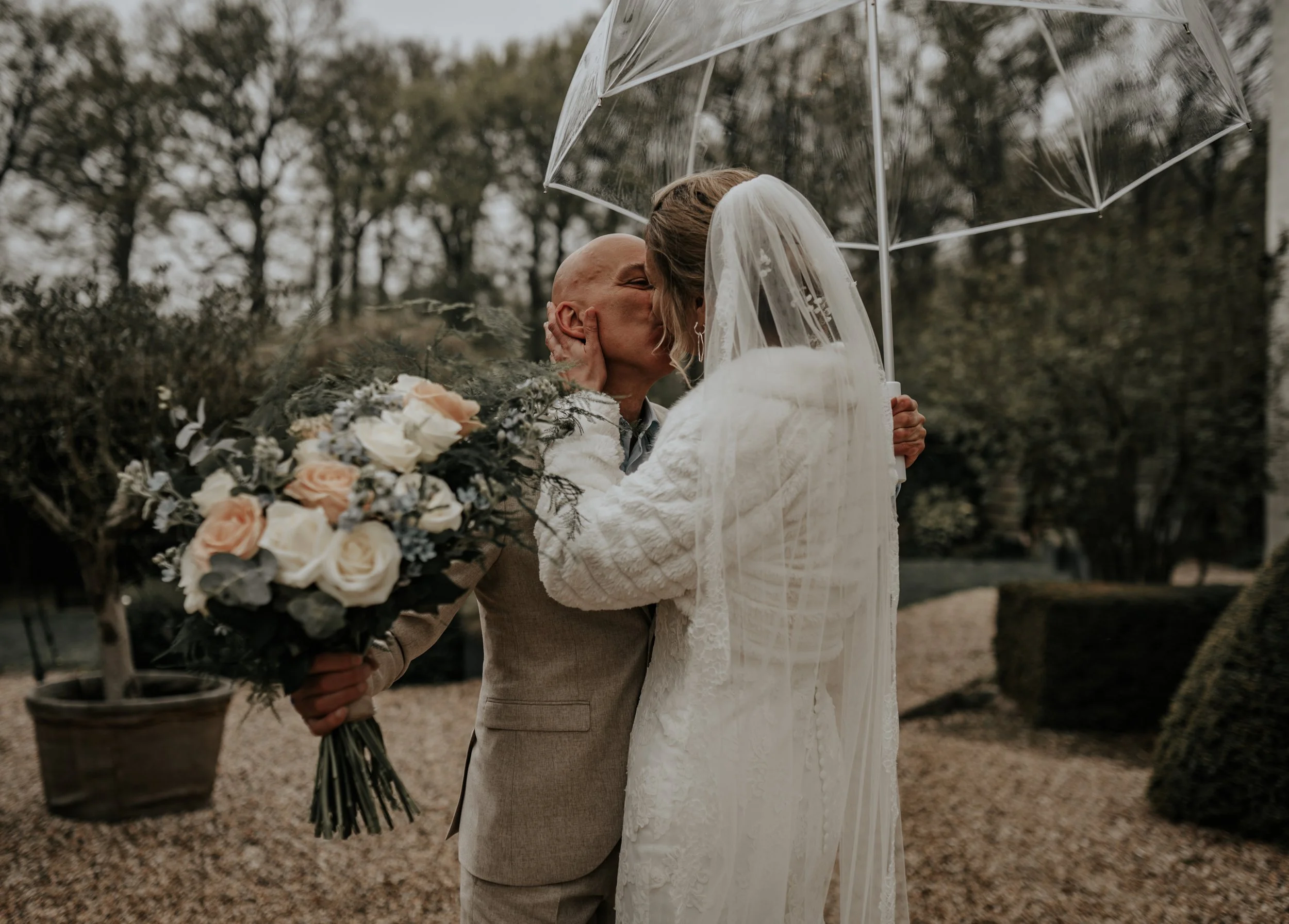 Twee mensen kussen onder een doorzichtige paraplu, een persoon houdt een grote bloemenschikking vast, op een buitenlocatie.