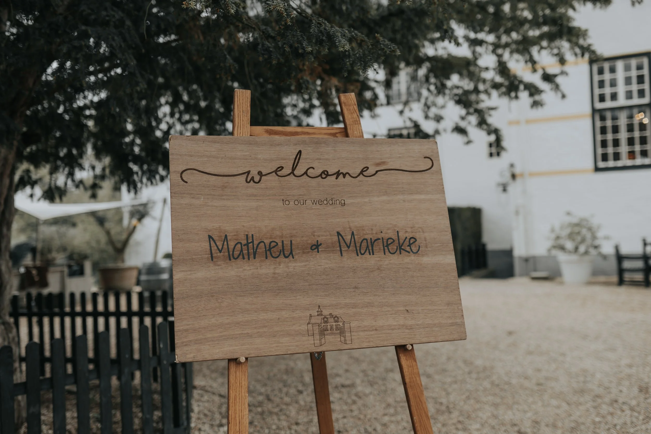 Een houten welkomstbord met tekst voor een bruiloft, vermeldt de namen Matheu en Marieke, geplaatst op een eikenhouten standaard buiten onder een boom, met een witte gebouw en tuin in de achtergrond.