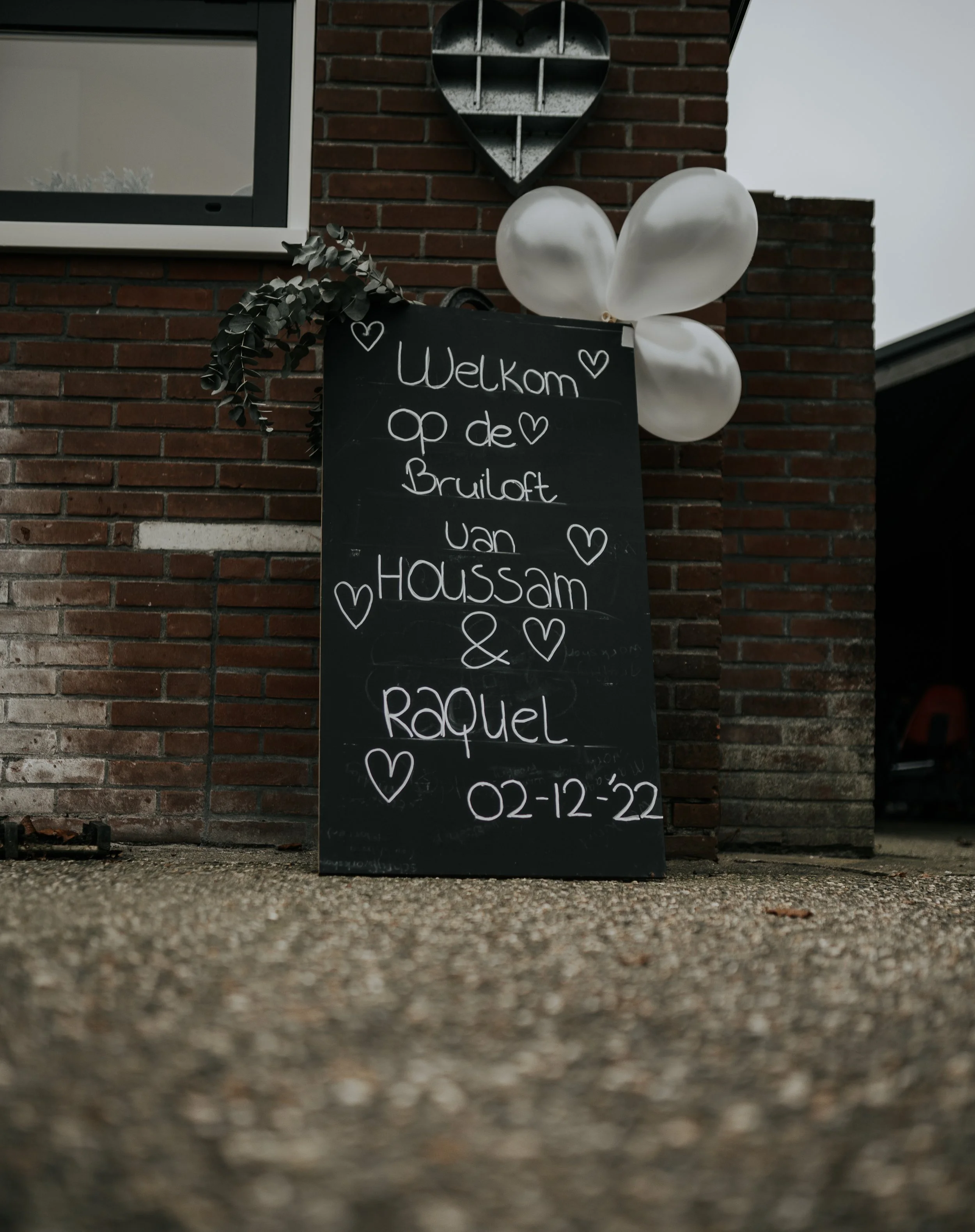 Een zwart bord met witte tekst die welkom zegt op de bruiloft van Haussam en Raquel, met ballonnen en hartversiering, afgesloten met de datum 02-12-22, geplaatst tegen een bakstenen muur.