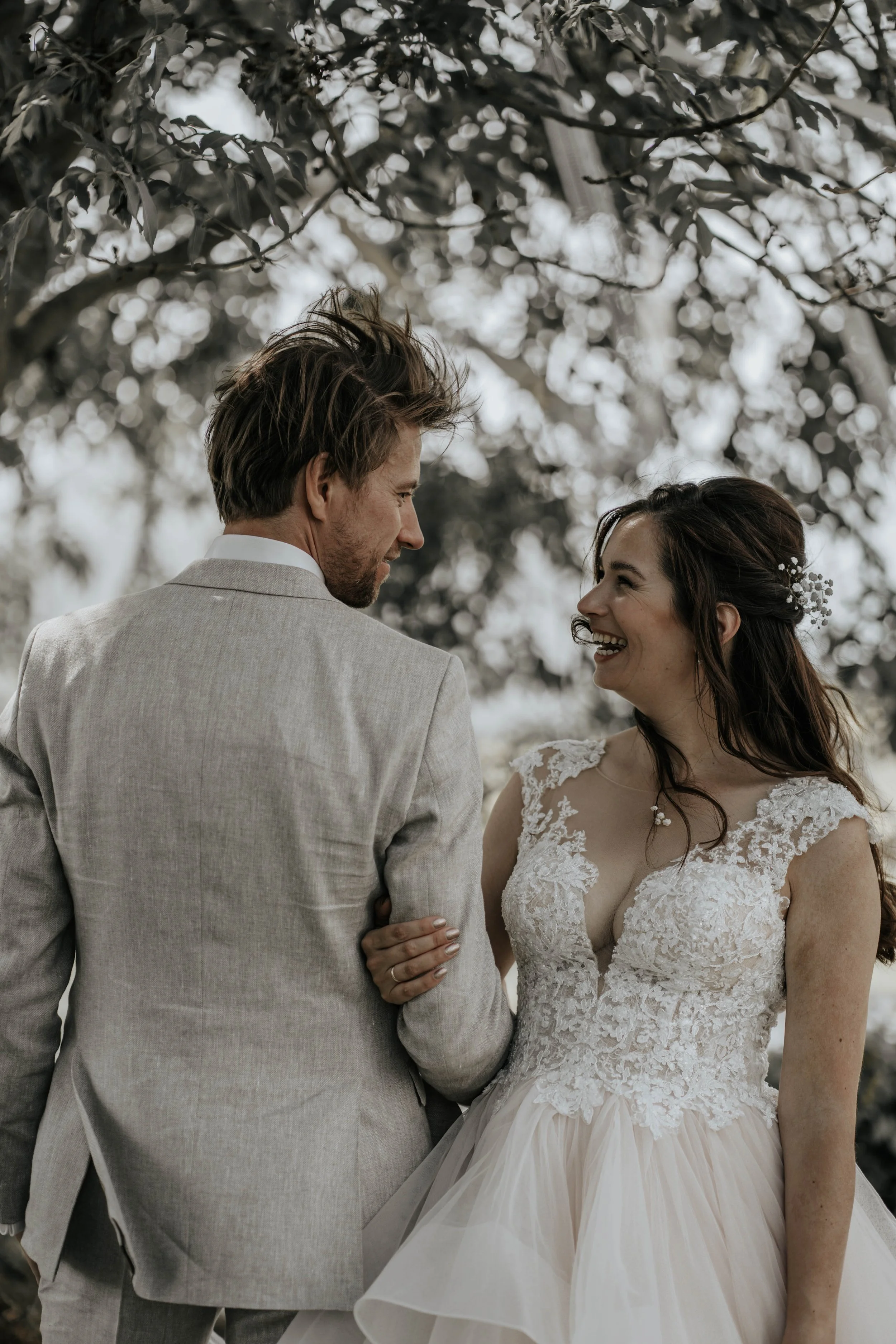 Een bruid en bruidegom die naar elkaar glimlachen onder een boom, op hun huwelijksdag, buiten.