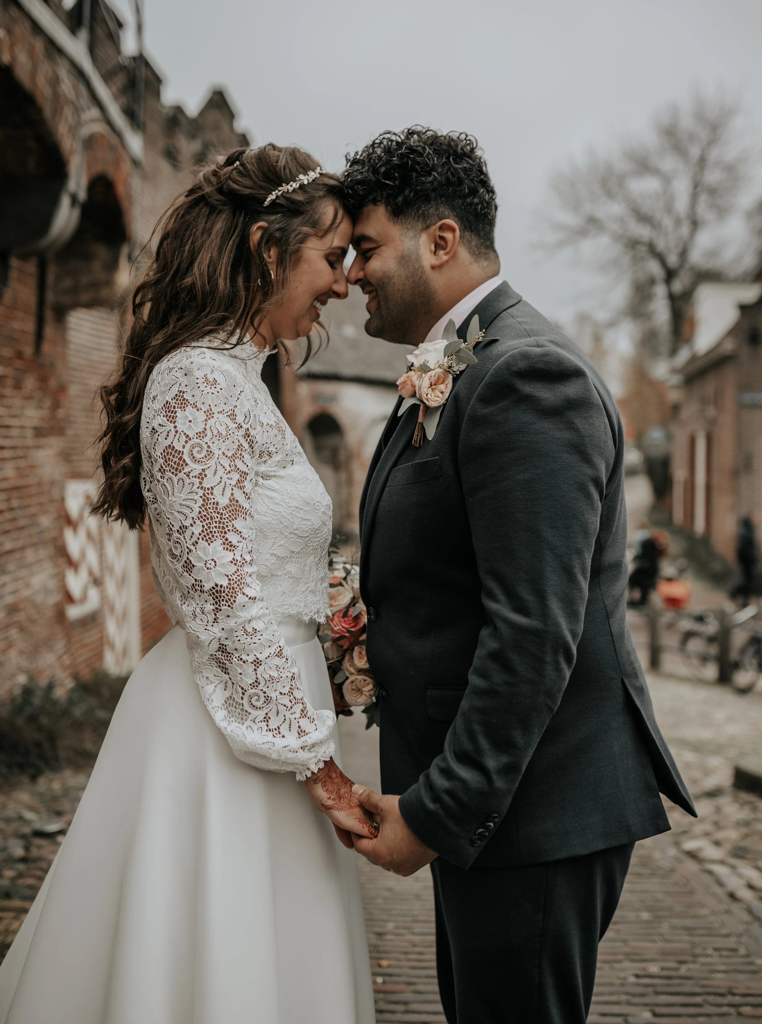 Een pas getrouwd stel dat dicht bij elkaar staat, hun voorhoofd tegen elkaar, in een straat met oude bakstenen gebouwen. Ze lachen en houden elkaars handen vast, de vrouw draagt een witte kanten jurk en de man een grijze pak met bloemen. Het is een herfstachtige dag met een grijze lucht.