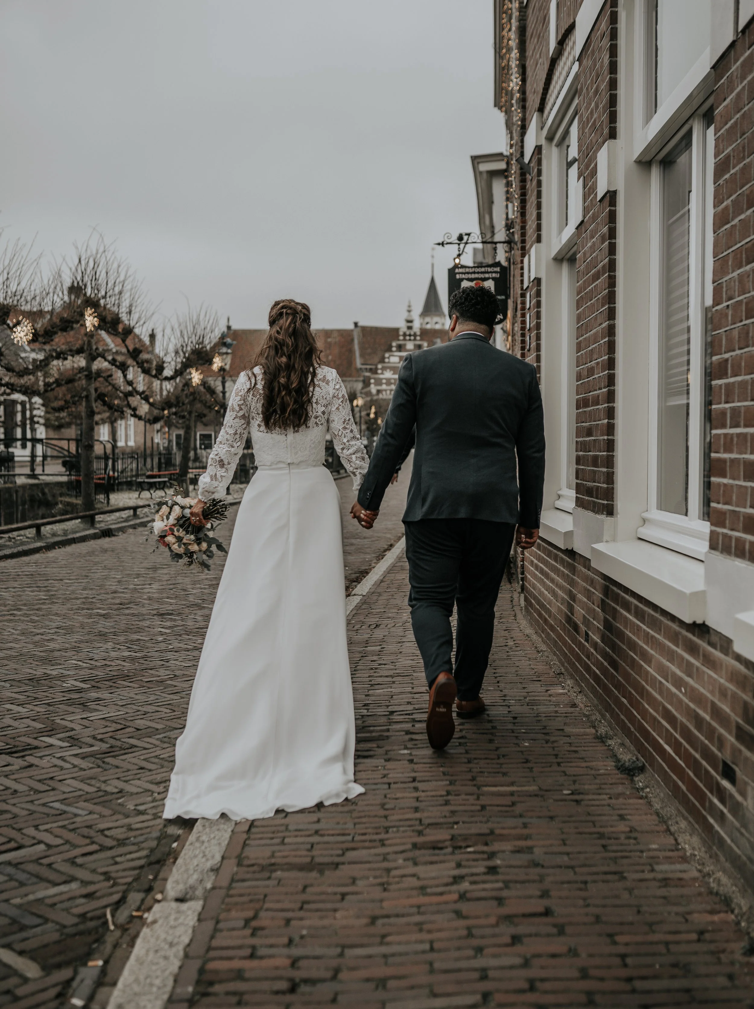 Een bruid en bruidegom lopen hand in hand over een straatje, de bruid draagt een witte trouwjurk met lange mouwen en heeft een bruidsboeket, de bruidegom draagt een donker pak. Het is een grijze dag, en de straat is versierd met kerstdecoraties.