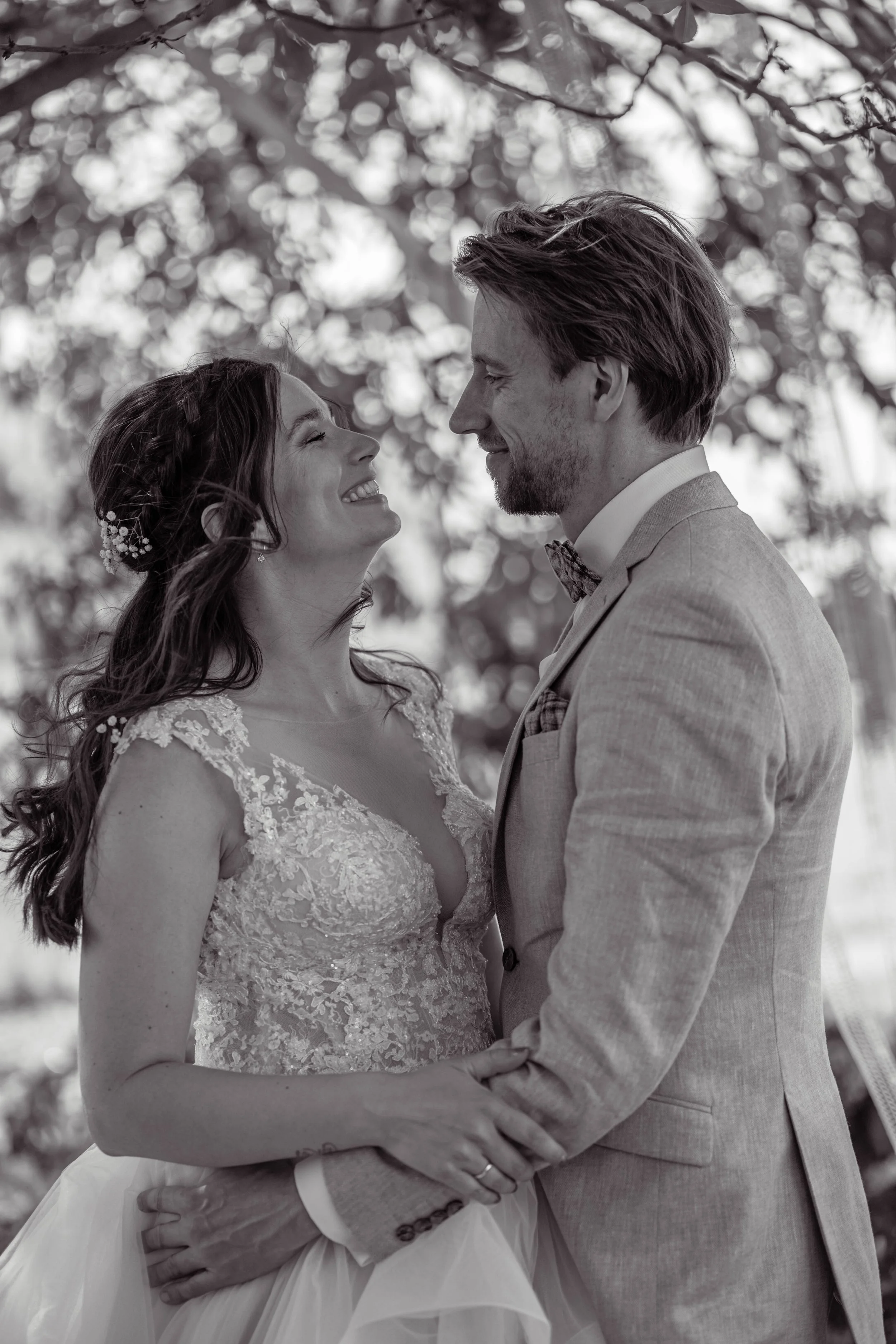 Een zwart-wit foto van een bruid en bruidegom die naar elkaar glimlachen en elkaar vasthouden, staan onder een boom.