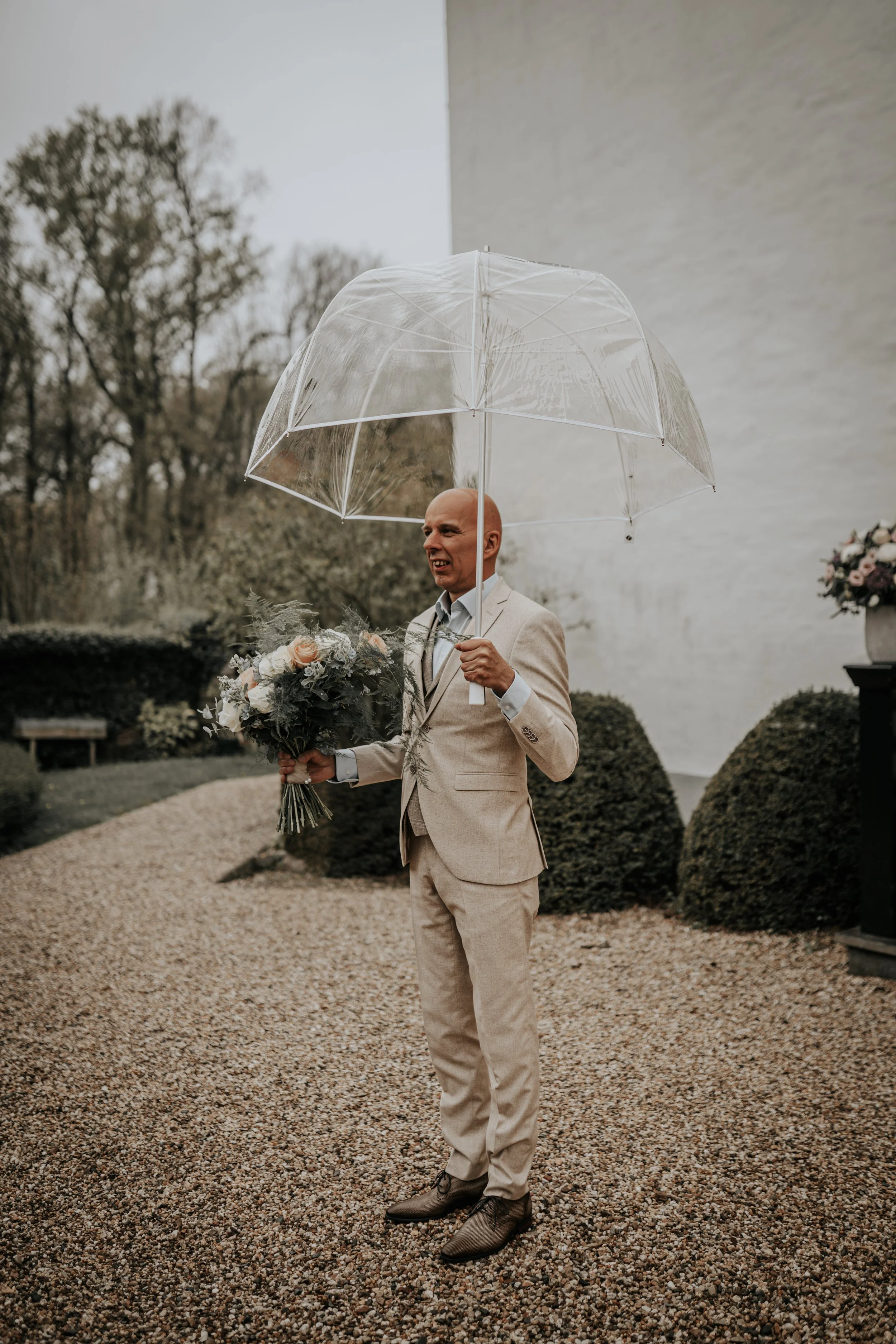 Man in een beige pak met bloemboeket onder een doorzichtige paraplu, buiten, op een gravelpad bij een witte muur en groen gebladerte.