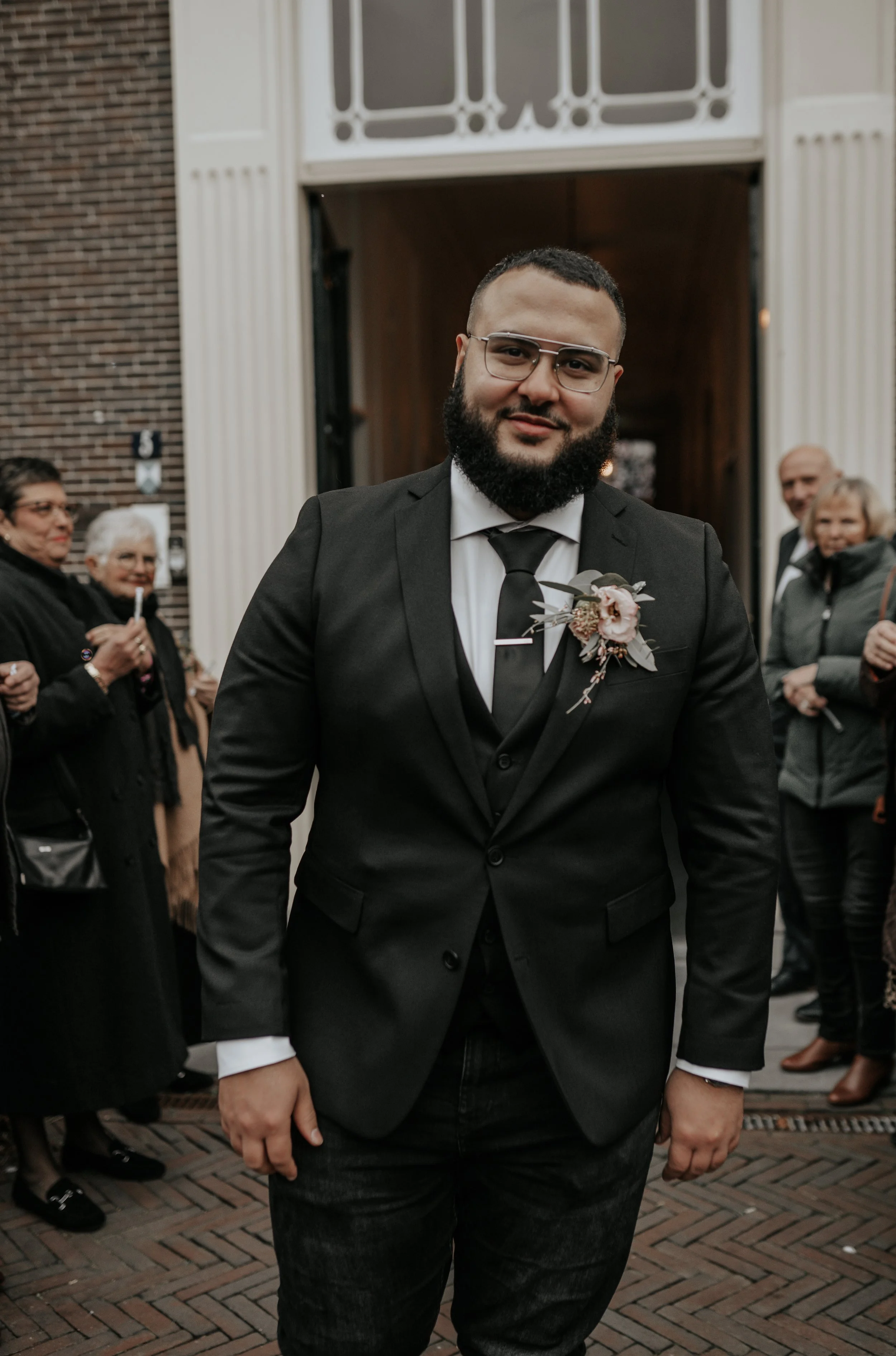 Man in een zwart pak met een bloem op de revers, omringd door andere mensen buiten voor een gebouw.