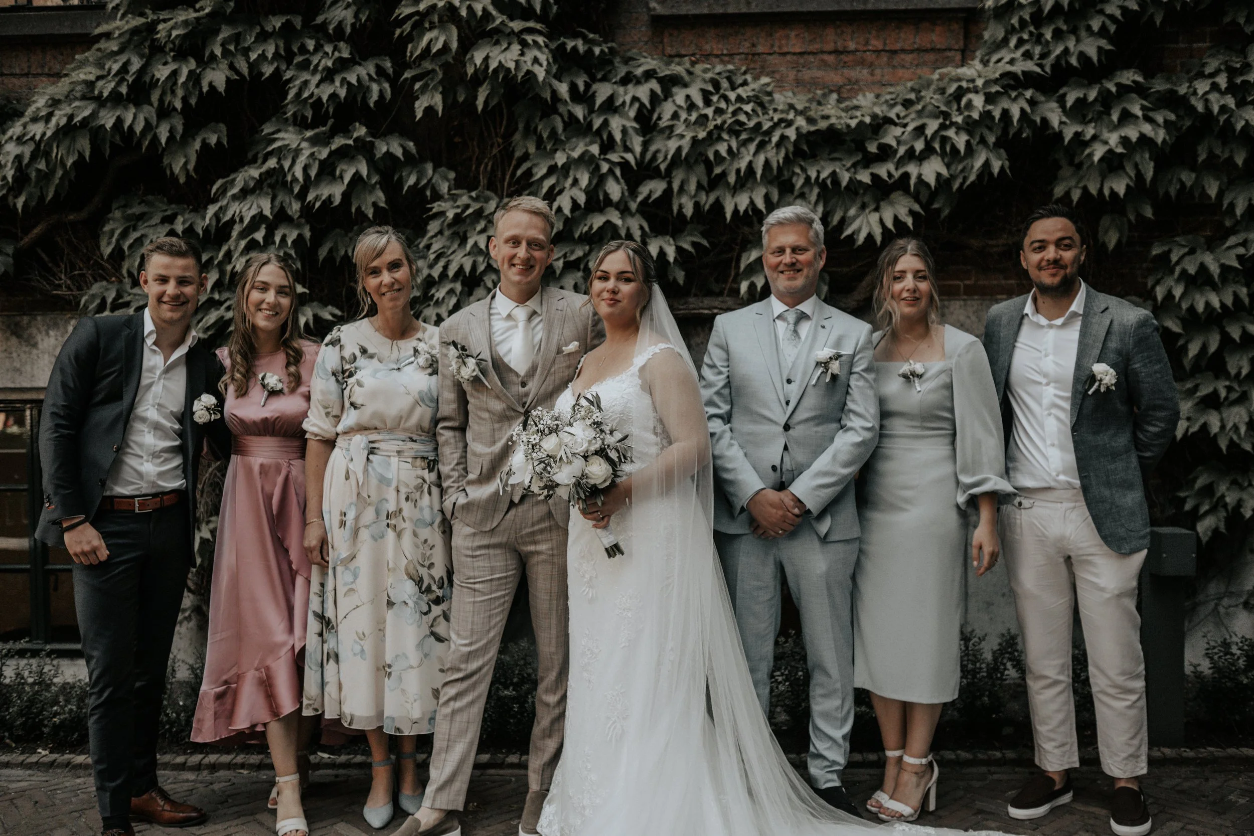 Groepsfoto van een bruiloft met acht mensen, waaronder een bruid en bruidegom, voor een muur bedekt met groene klimplanten.