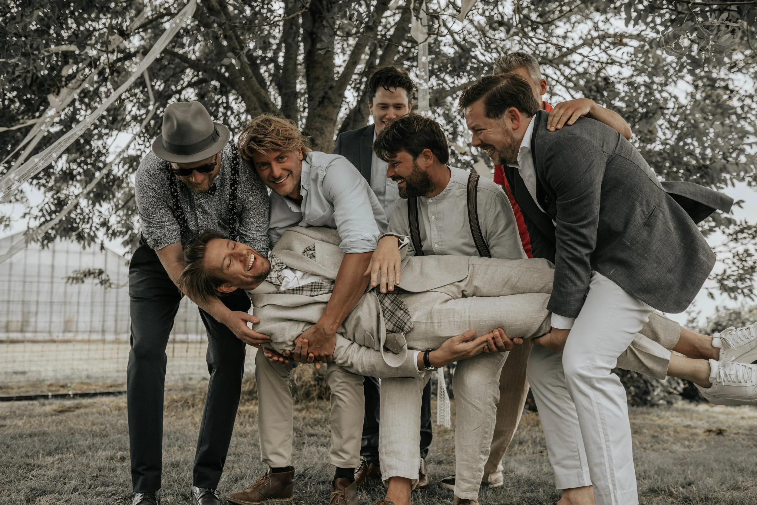 Een groep mannen tilt iemand die doet alsof hij ziek is in een parkwandeling, allen glimlachend en lachend op een grasveld onder een grote boom.