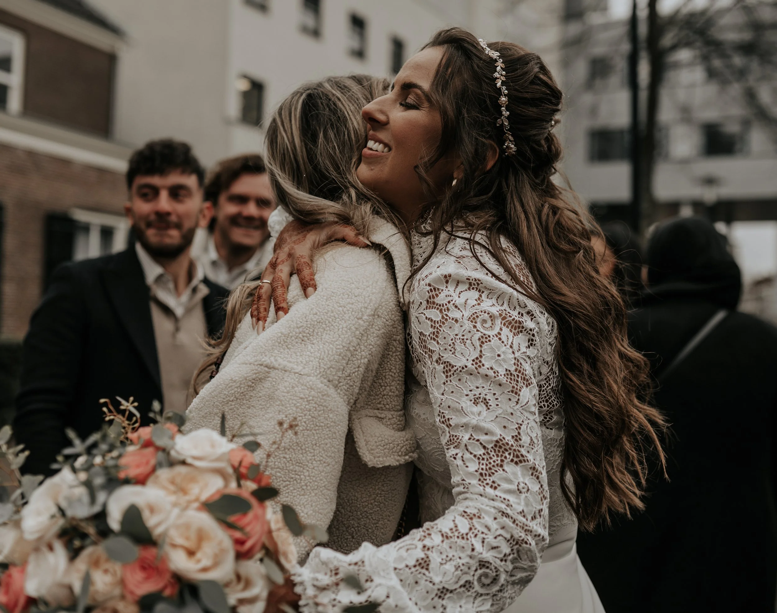 Man en vrouw omhelzen, omringd door lachende mensen, buiten, bruiloft, vrouw in kanten jurk met bloemaccessoire in haar haar, vrouw houdt een boeket bloemen.