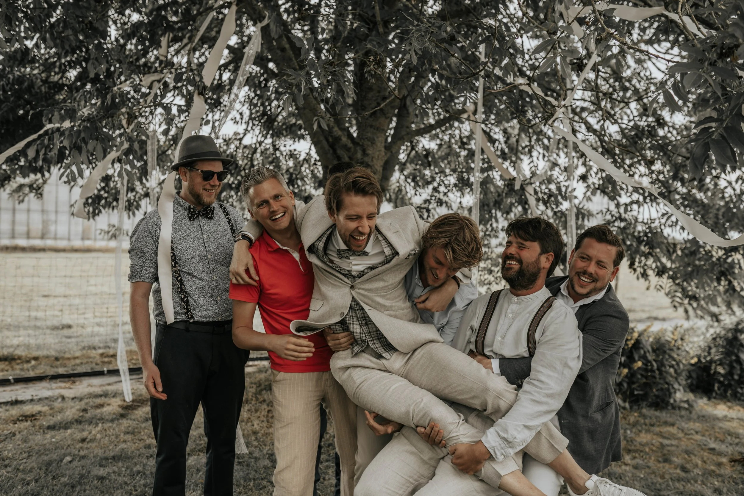 Vijf mannen lachen en spelen samen onder een boom, één tilgt een ander op terwijl de anderen hem ondersteunen, tijdens een feest of evenement in de buitenlucht.