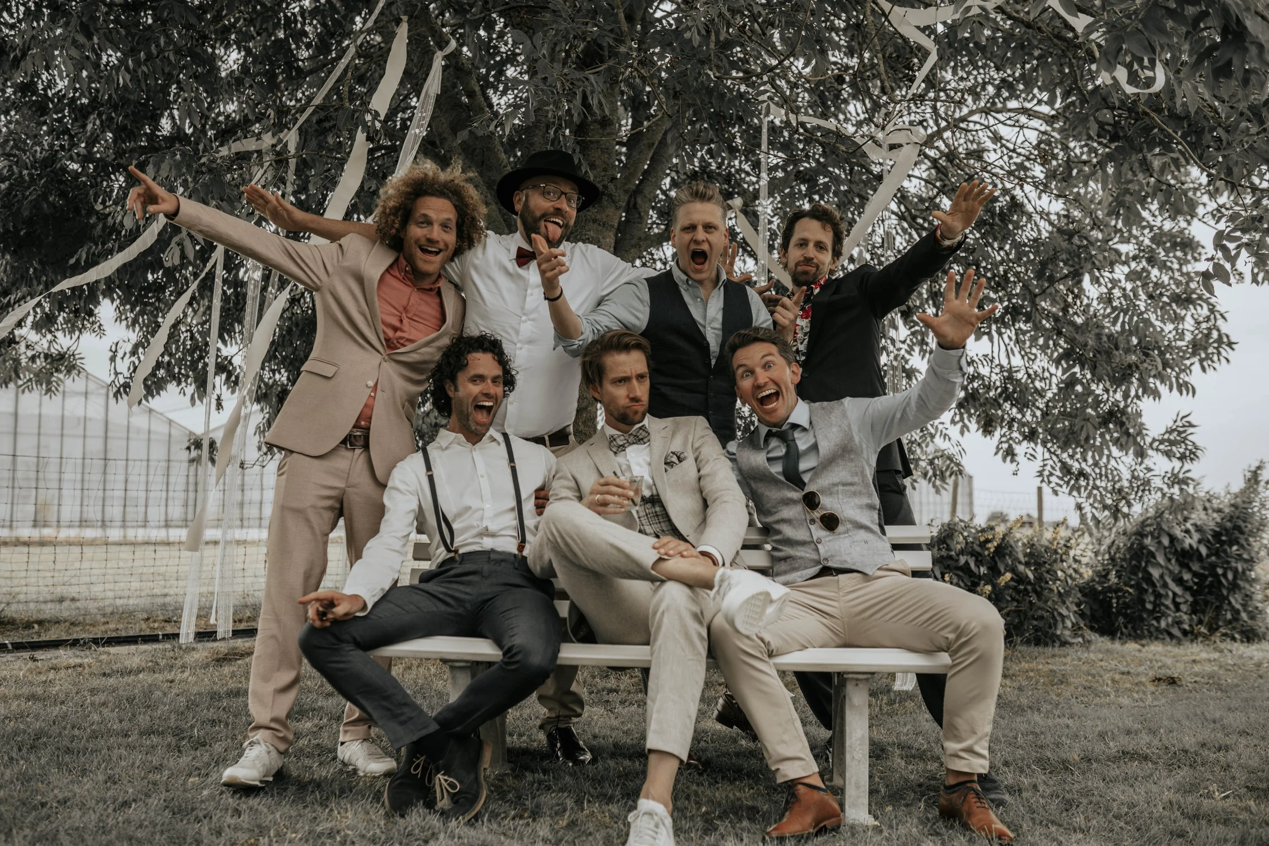 Een groep mannen in formele en casual kleding die buiten onder een boom op een bank zitten en poseren voor de camera, sommigen lachen en maken grappige gezichten.