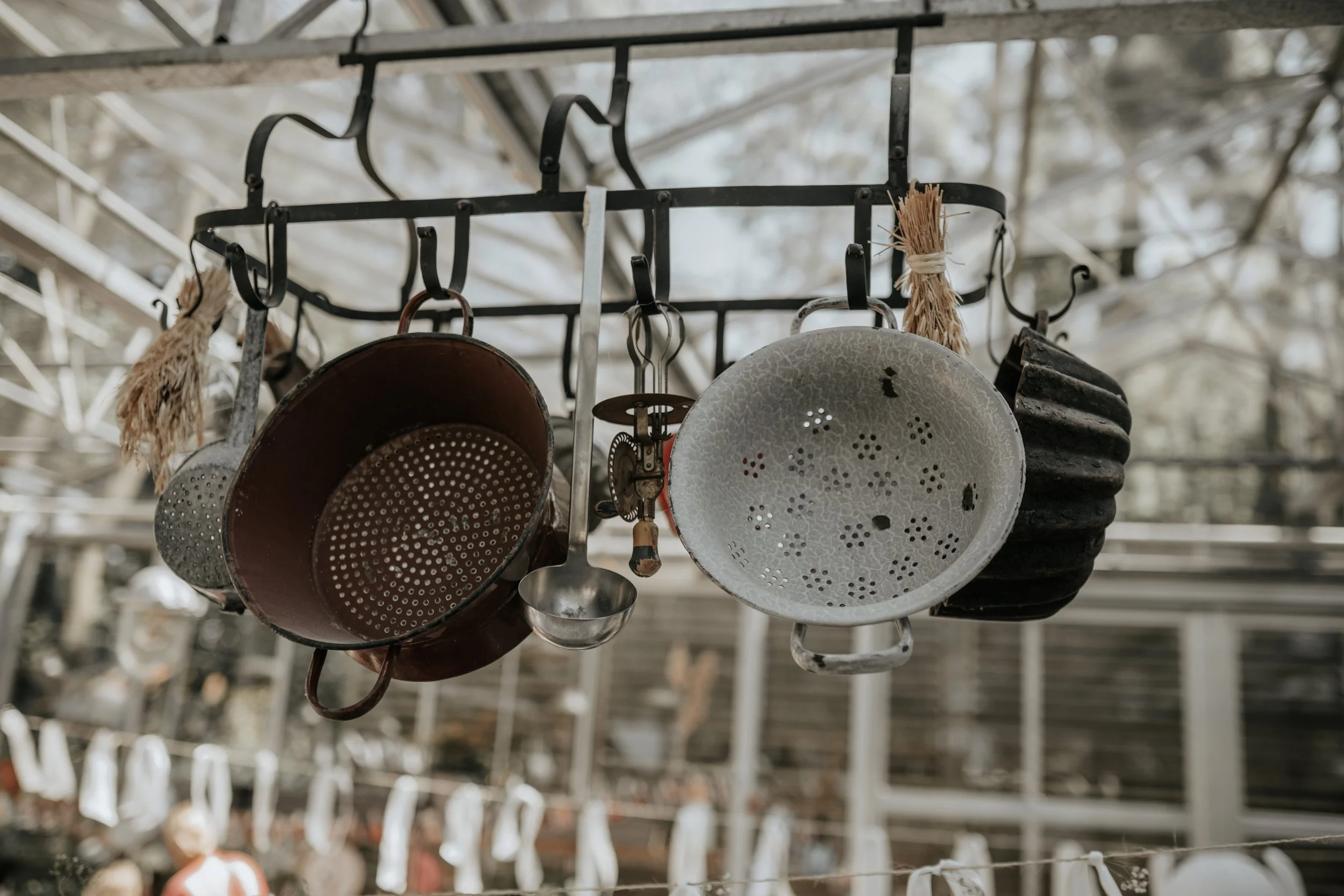 Vijf oude metalen keukenkruiken en pannen hangen aan een hakenrek buiten.