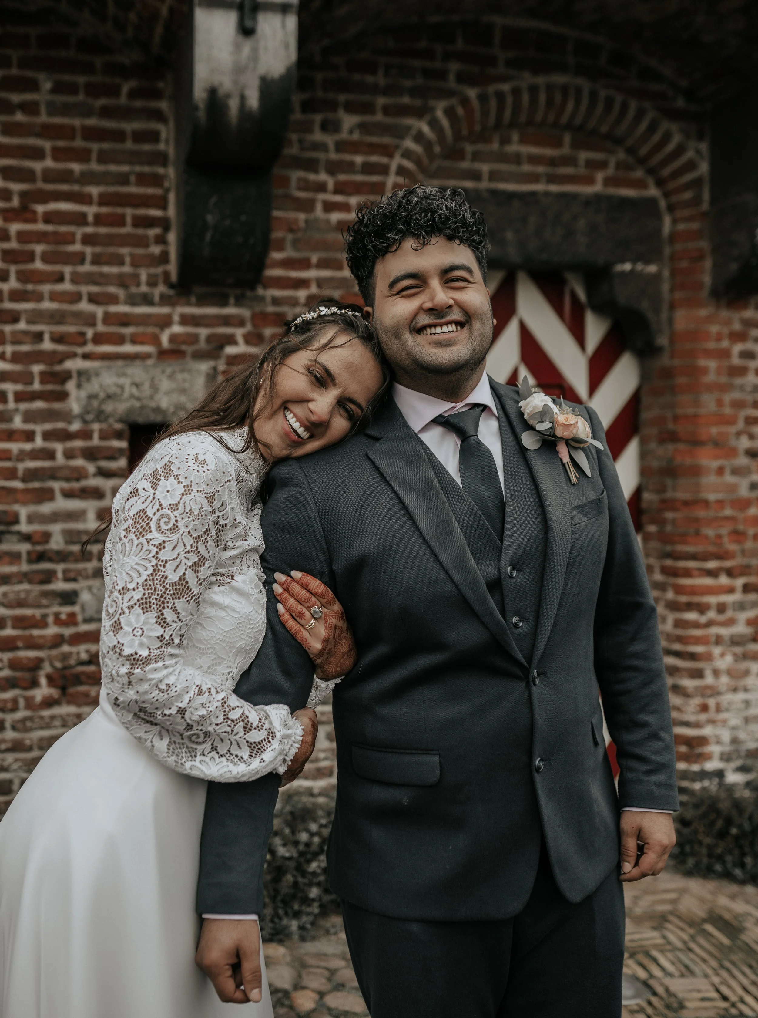 Een bruid en bruidegom glimlachen op hun trouwdag voor een bakstenen muur.