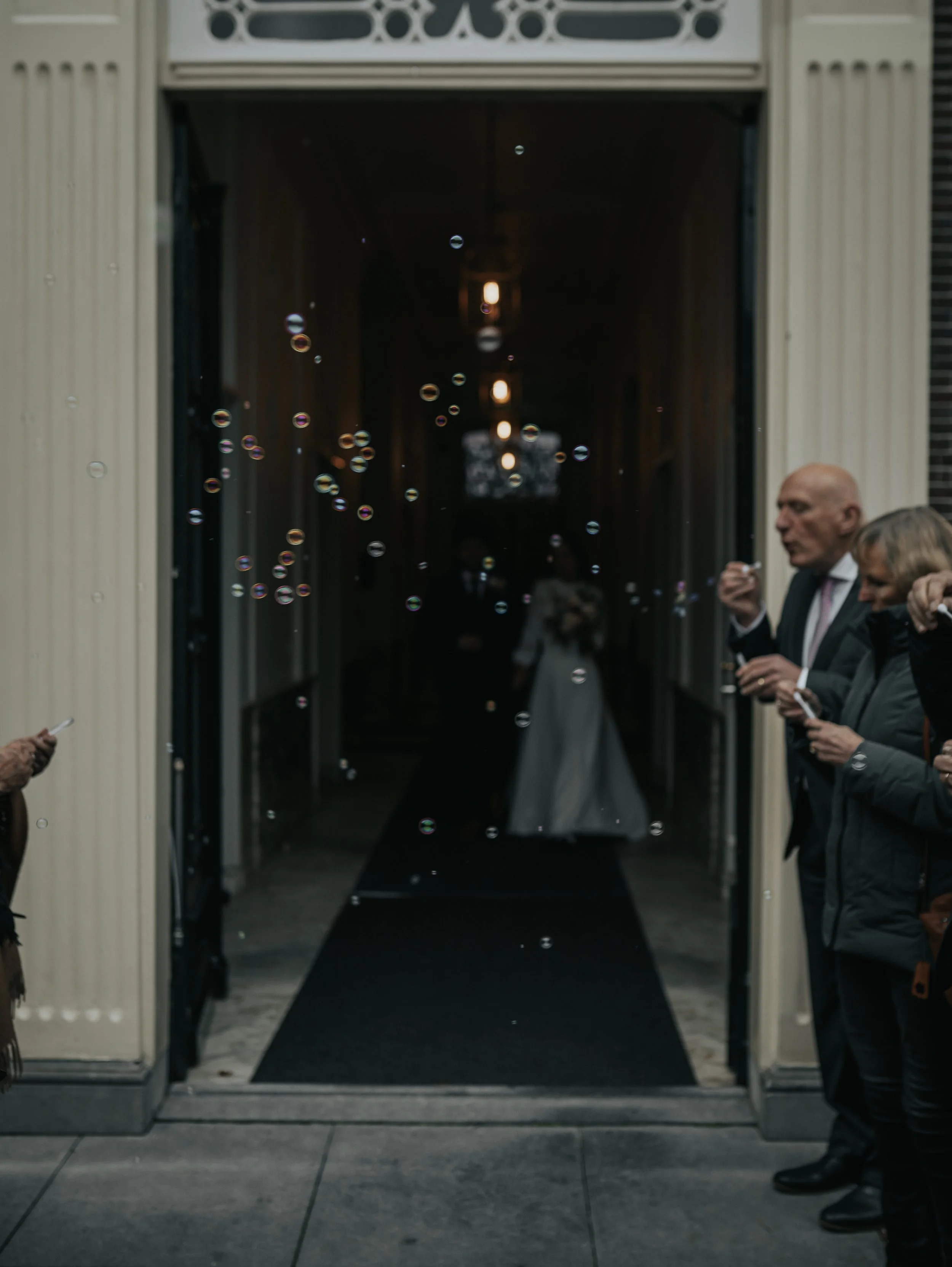 Gasten wachten buiten bij de ingang van een gebouw voor een bruiloft, met enkele bellen die in de lucht zweven.