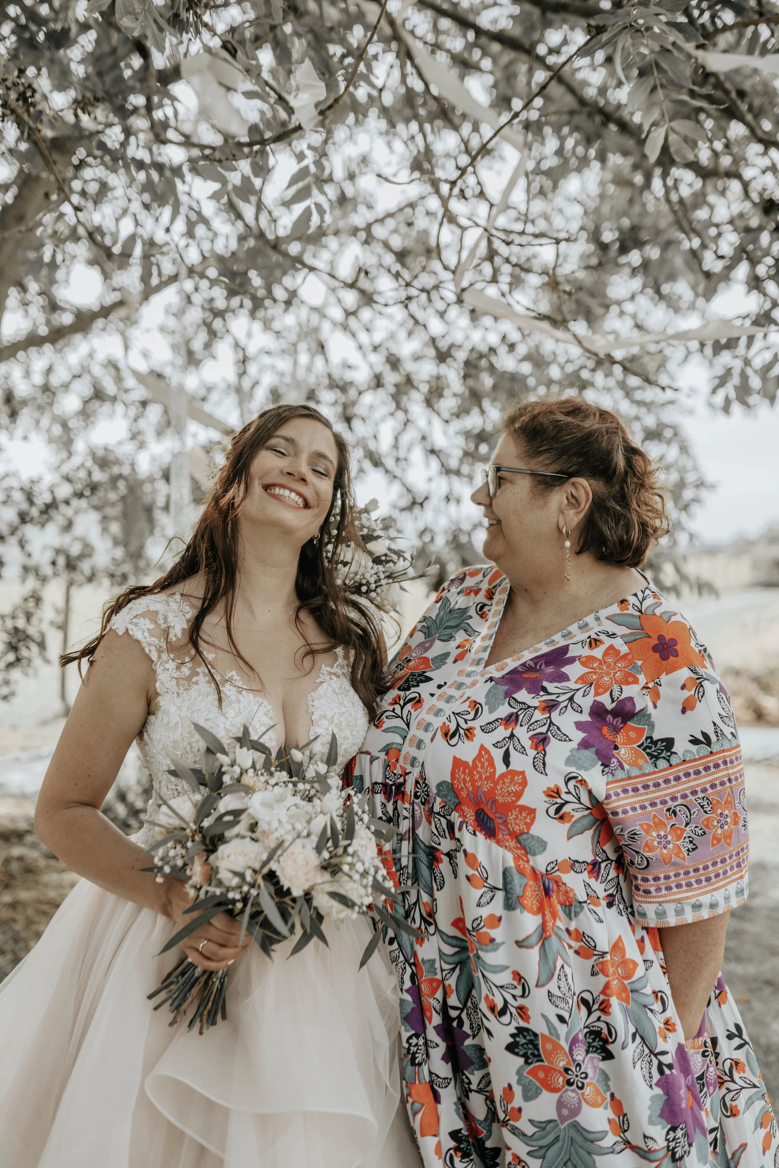 Twee vrouwen, een bruid in een witte jurk en een vrouw in een kleurrijk blouse, staan onder een boom met witte bloemen, glimlachend en lachend naar elkaar.