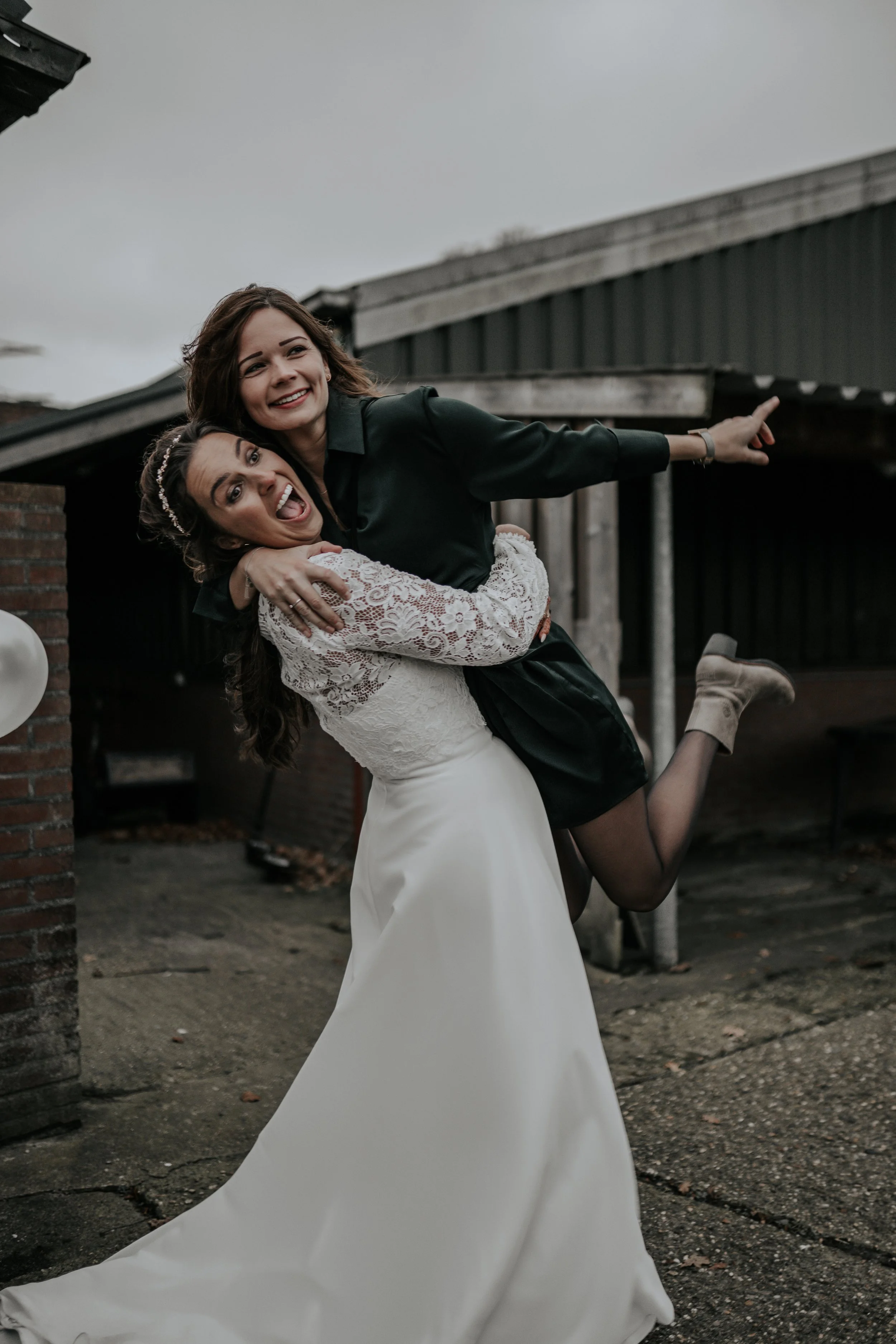 Twee vrouwen tijdens een bruiloft, één in een trouwjurk en de ander in een zwarte outfit, lachen en genieten buiten op een sombere dag.