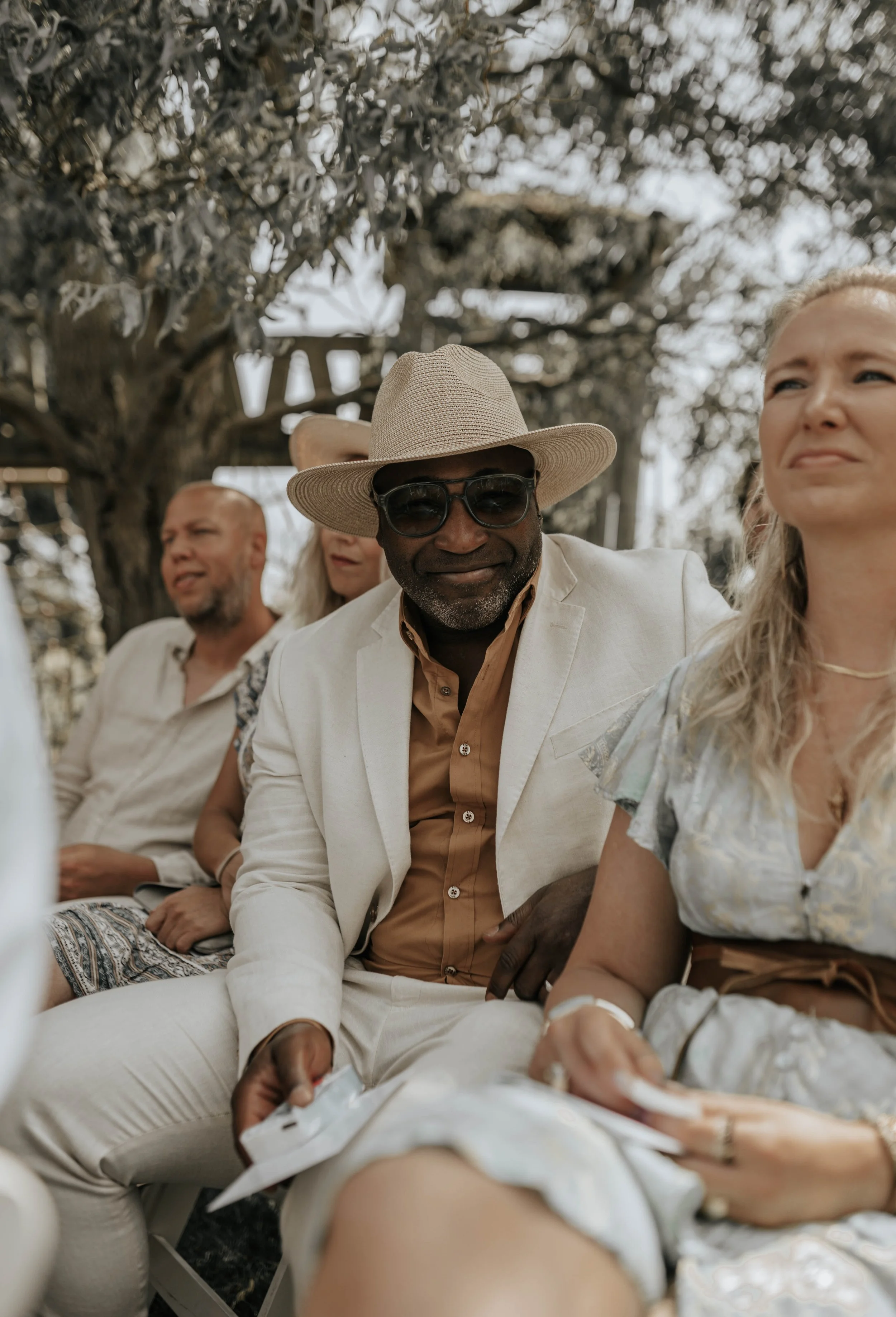 Mensen zitten buiten onder een boom, gekleed in zomerse kleding, met één man die een hoed en sunglasses draagt, mogelijk tijdens een evenement of bijeenkomst.