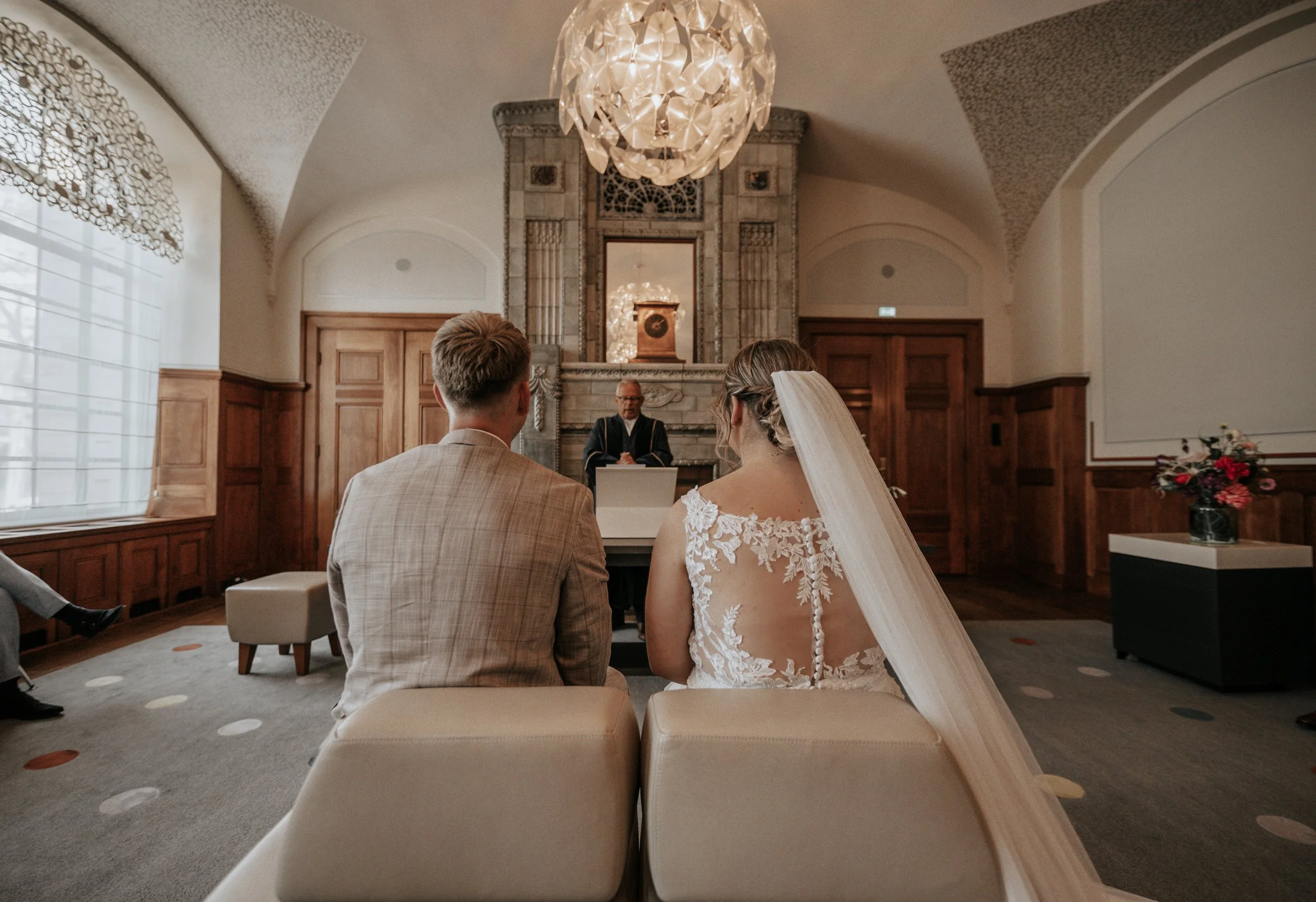 Bruidegom en bruid zitten samen tijdens een huwelijkceremonie in een elegante zaal met een open haard en een grote chandelier, terwijl een officiant voor hen spreekt.