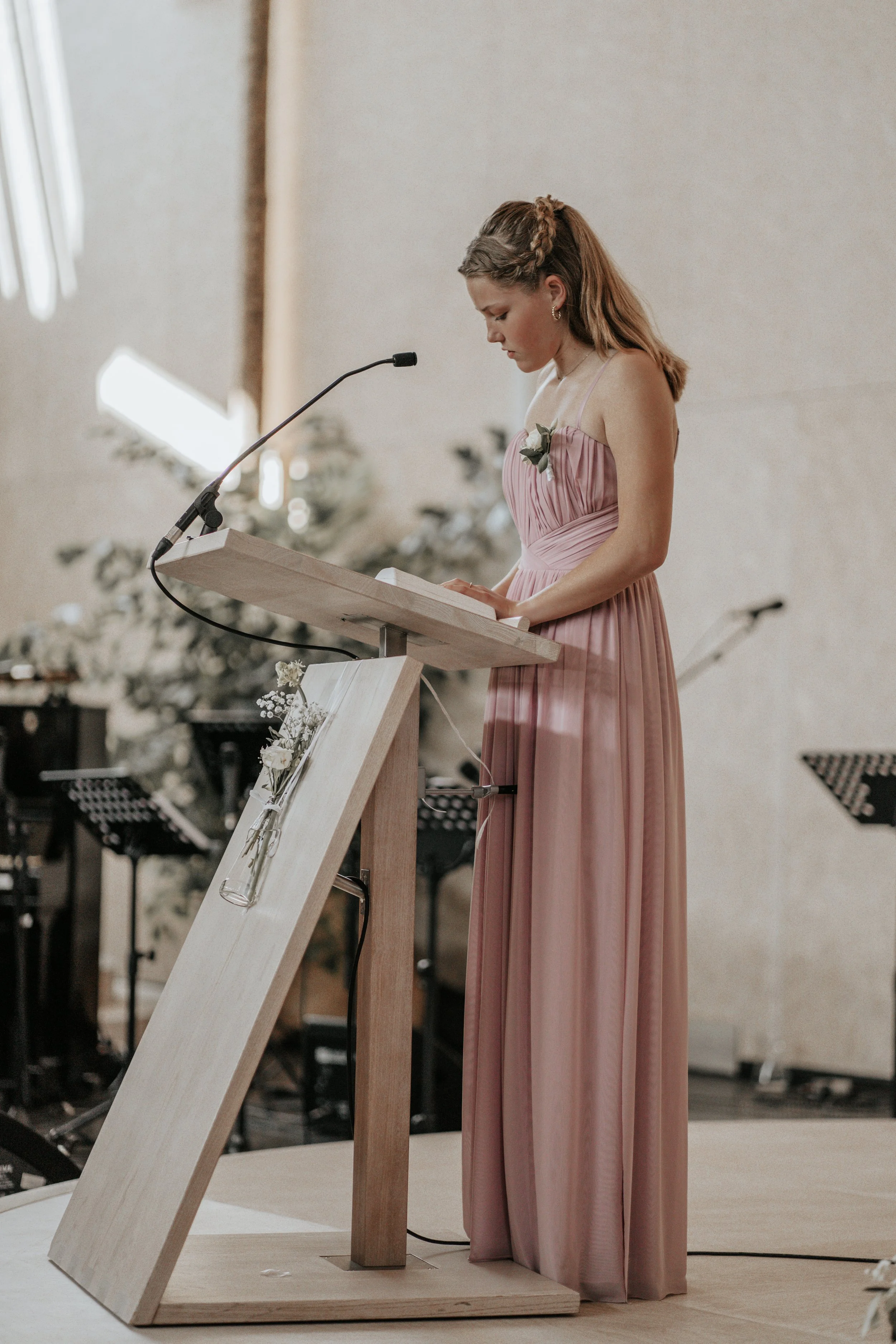 Een jonge vrouw in een lange, roze jurk staat achter een houten lessenaar en leest of spreekt in een microfoon tijdens een ceremonie, met bloemen en muziekinstrumenten op de achtergrond.