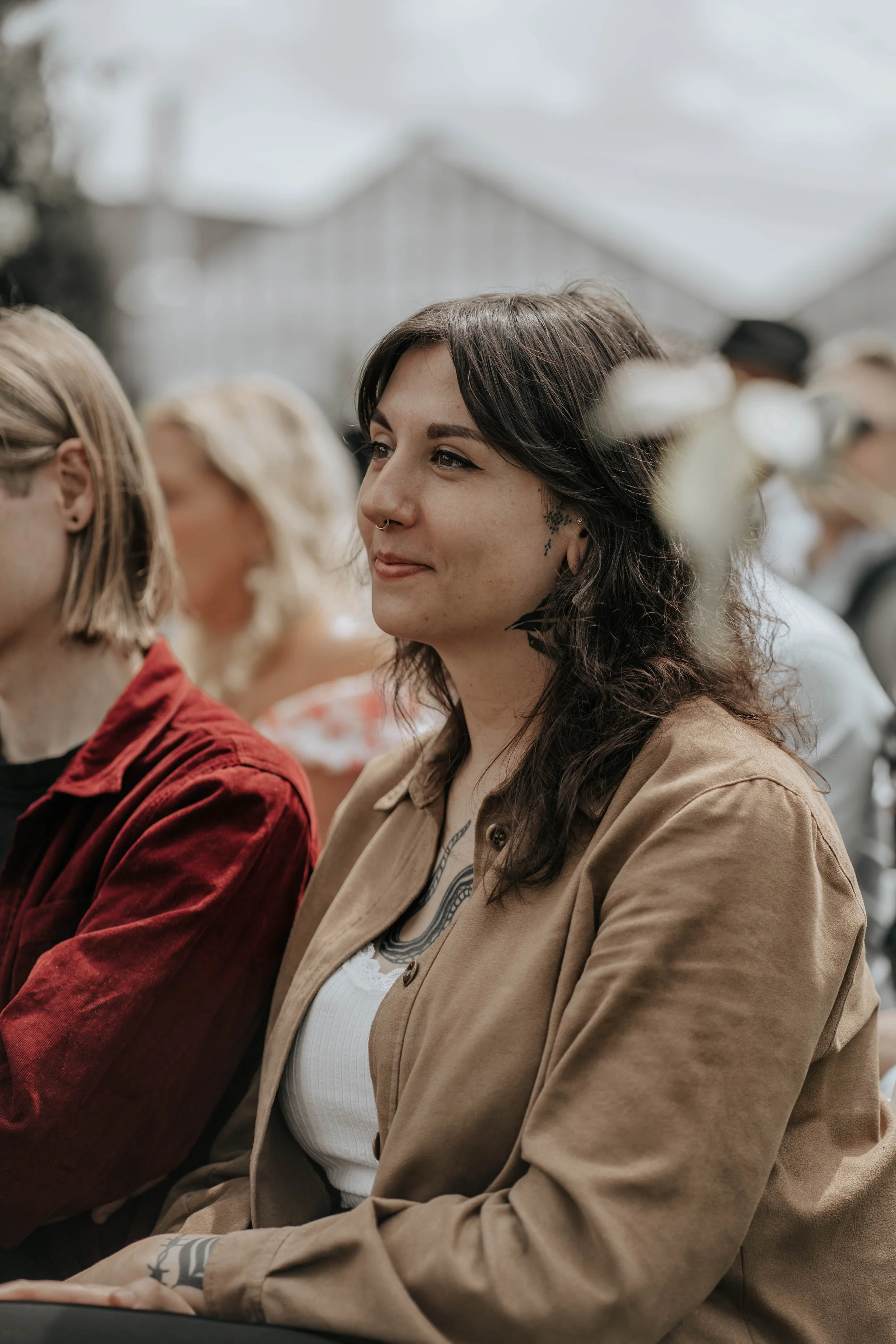 Tien mensen zitten naast elkaar in een semi-outdoor setting, de vrouw in het midden heeft donker, golvend haar en draagt een beige jas en een witte top, ze kijkt tevreden.