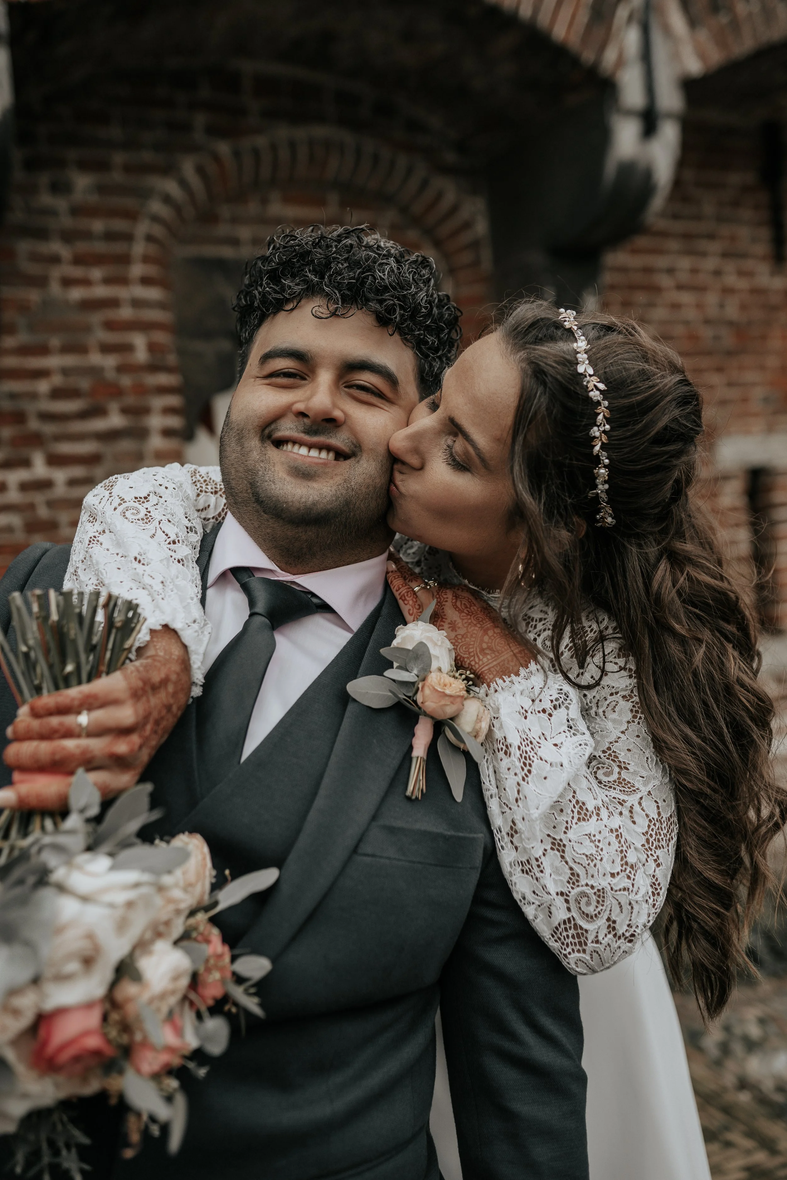 Bruidegom met bruid die hem kust, ze is in een witte kanten jurk en heeft een diadeem in haar haar, ze omarmen elkaar liefdevol buiten een bakstenen muur.