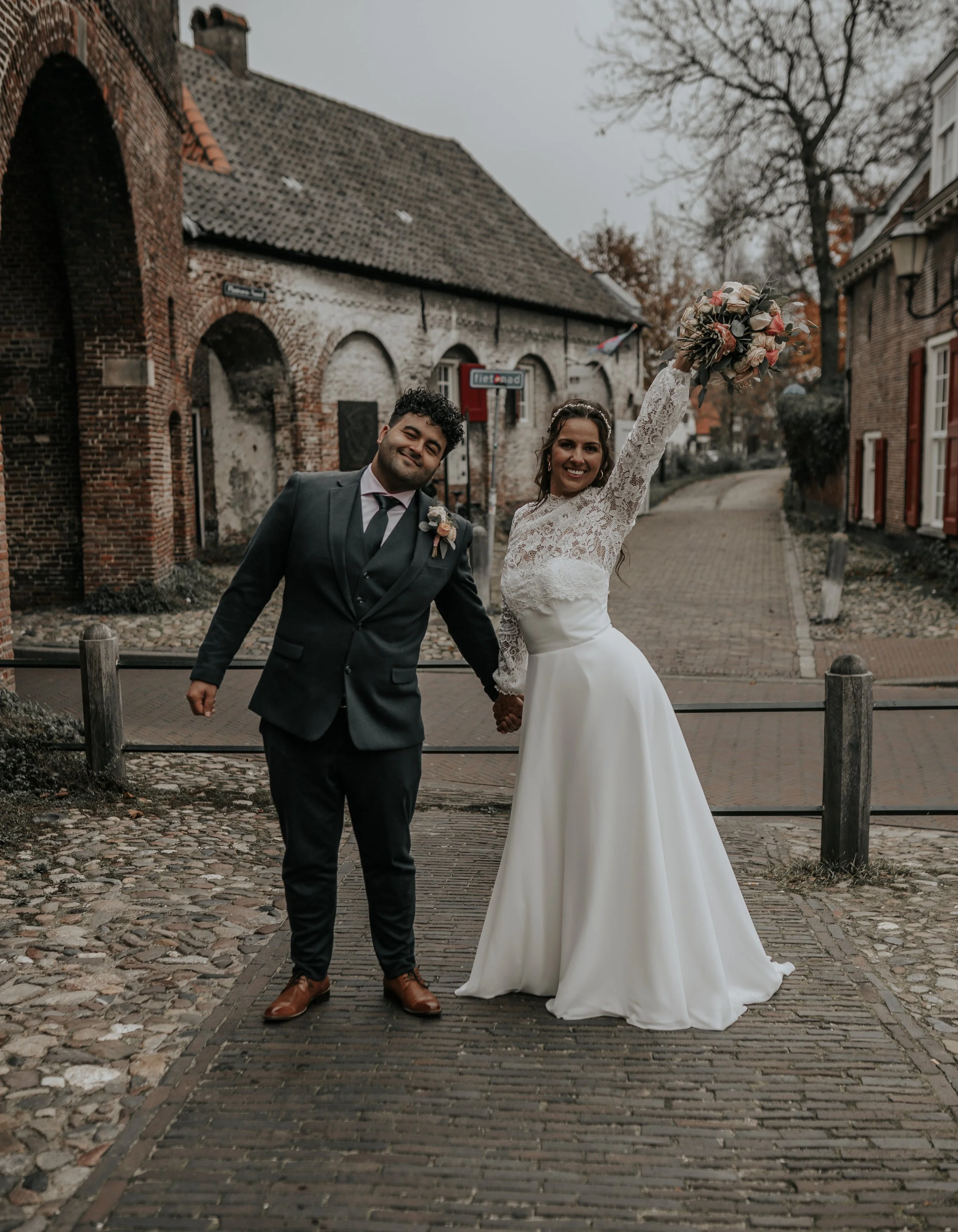 Een blije bruid en bruidegom die hand in hand staan op een straat met stenen en klinkers. De bruid houdt haar arm omhoog met een bloemenboeket. Het weer is grijs en de bomen op de achtergrond zijn kaal.