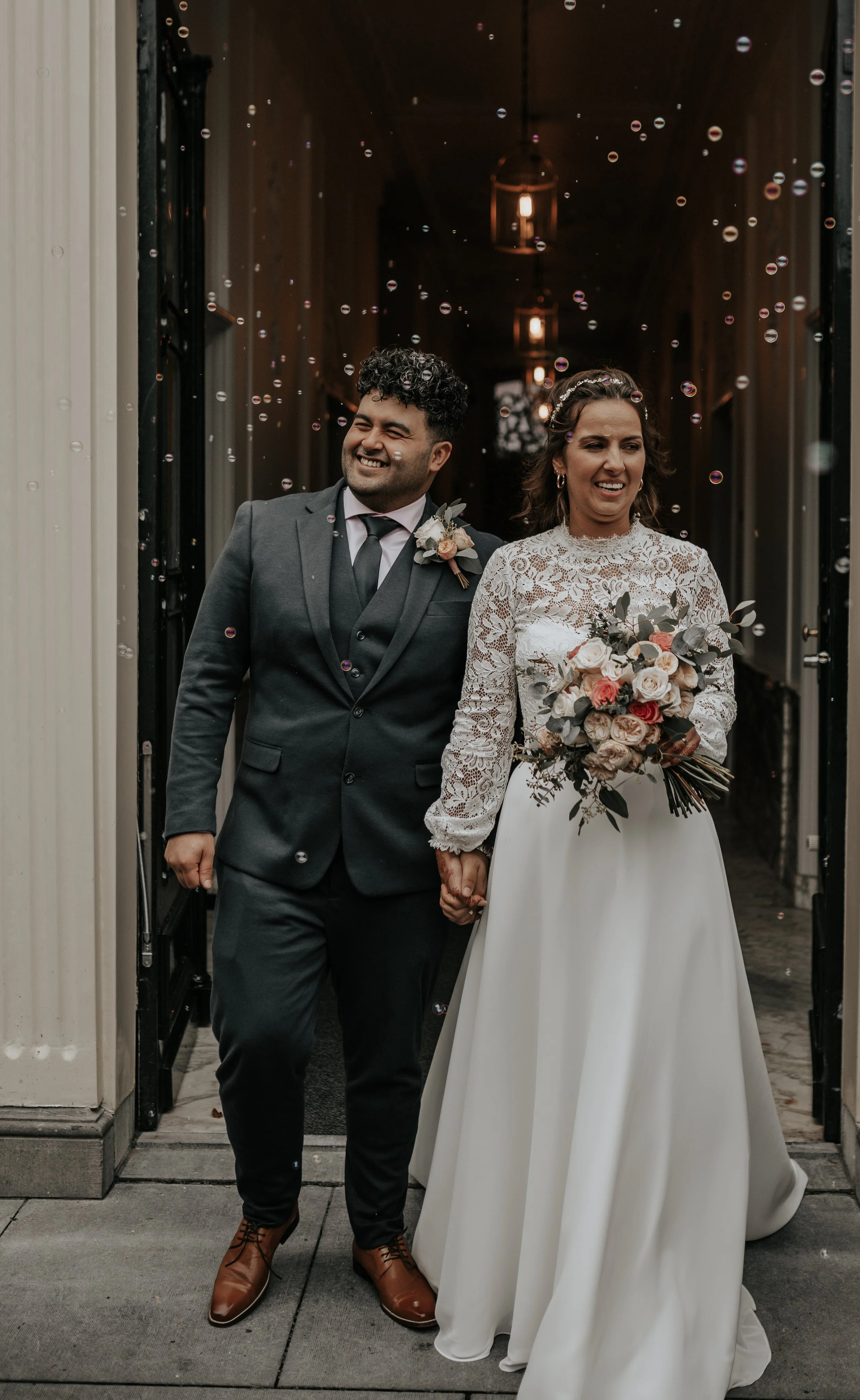 Een stel op hun bruiloft die hand in hand naar buiten komen, omringd door bellen. De man draagt een donkergrijs pak met een wit overhemd en een zwarte stropdas, de vrouw draagt een witte trouwjurk met kant en houdt een bouquet van bloemen.