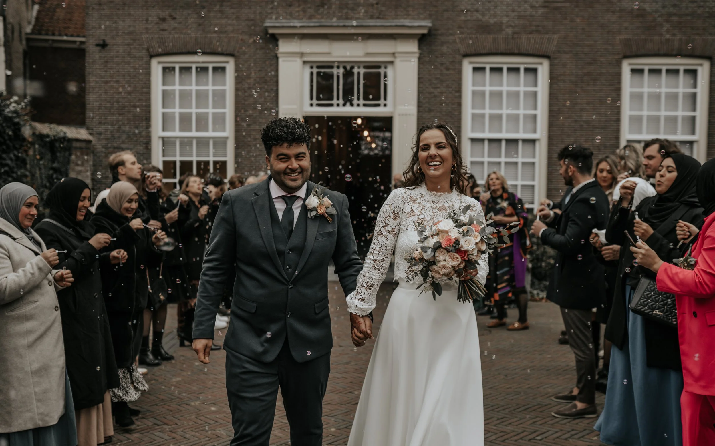 Een pasgetrouwd stel loopt hand in hand weg uit een feestelijke bijeenkomst, omringd door vrienden en familie die confetti en bellen blazen, buiten voor een groot gebouw.