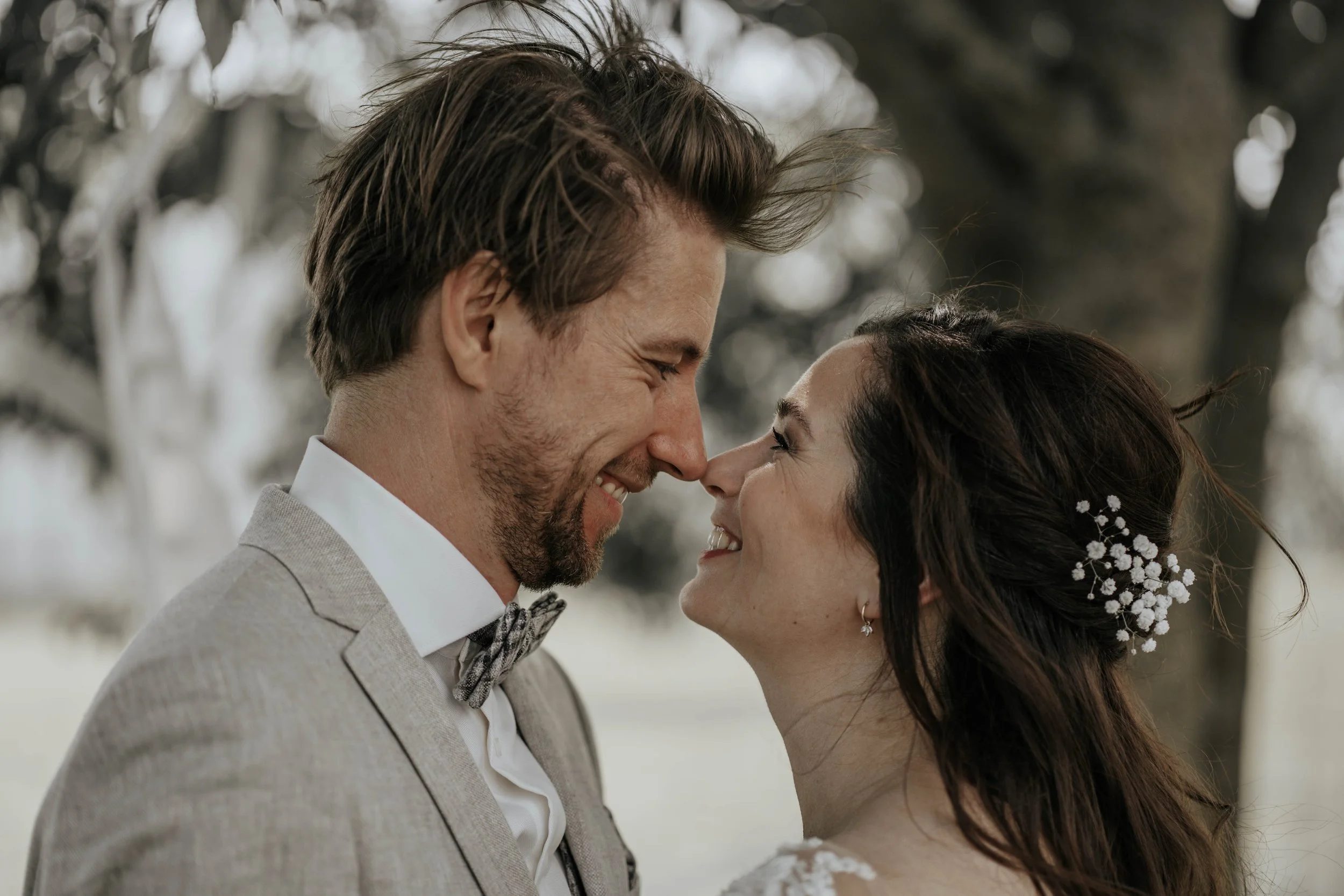 Een gelukkig koppel op hun bruiloft, die dicht tegen elkaar aan staan met voorhoofd tegen voorhoofd, lachend en met ogen dicht, buiten onder een boom.