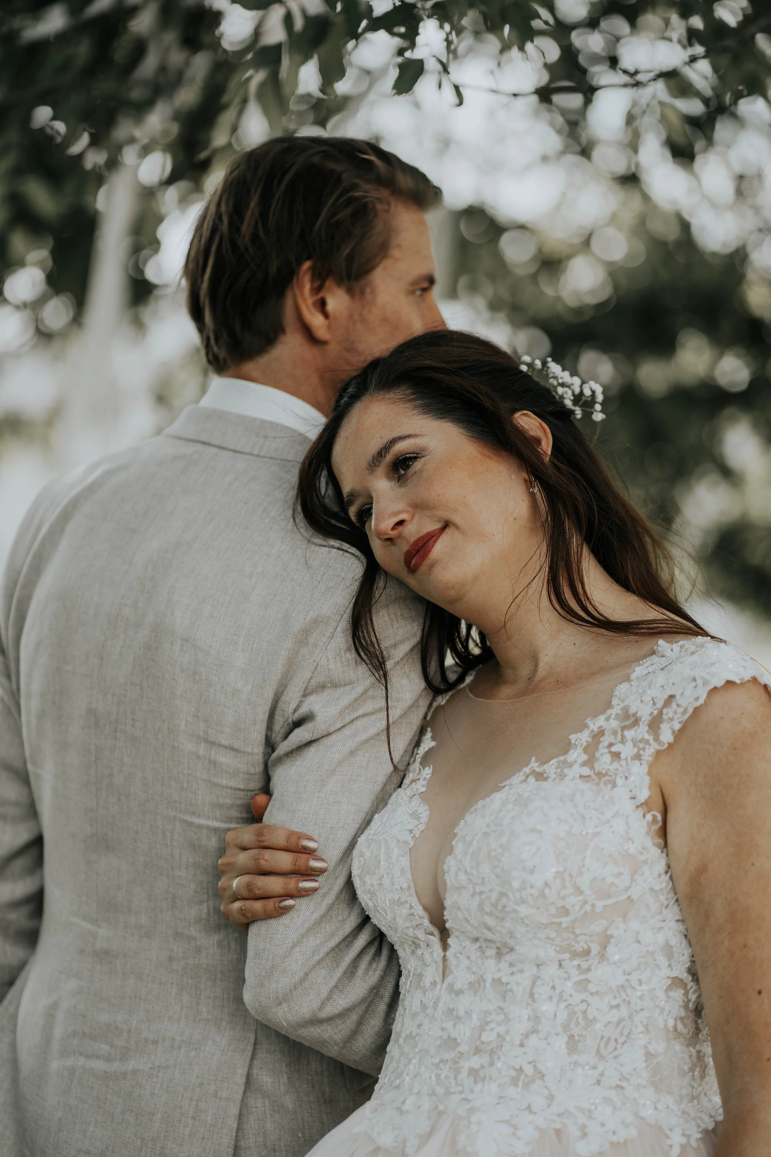 Een bruid en bruidegom omhelzen elkaar op een buitenceremonie, omringd door groene bladeren en bomen.