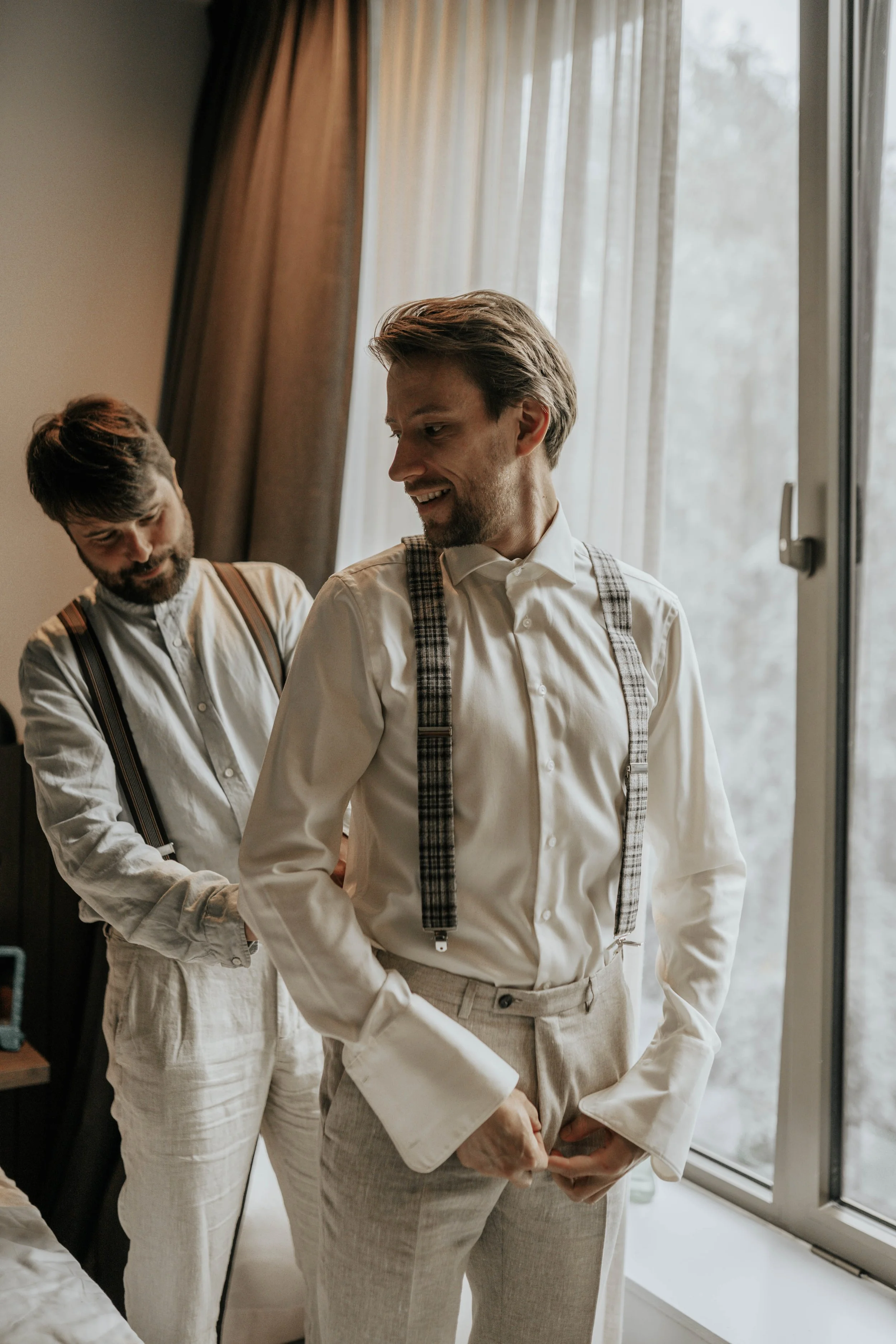 Twee mannen in netjes geklede kleding, voorbereidend voor een evenement of bruiloft, bij een raam in een kamer.