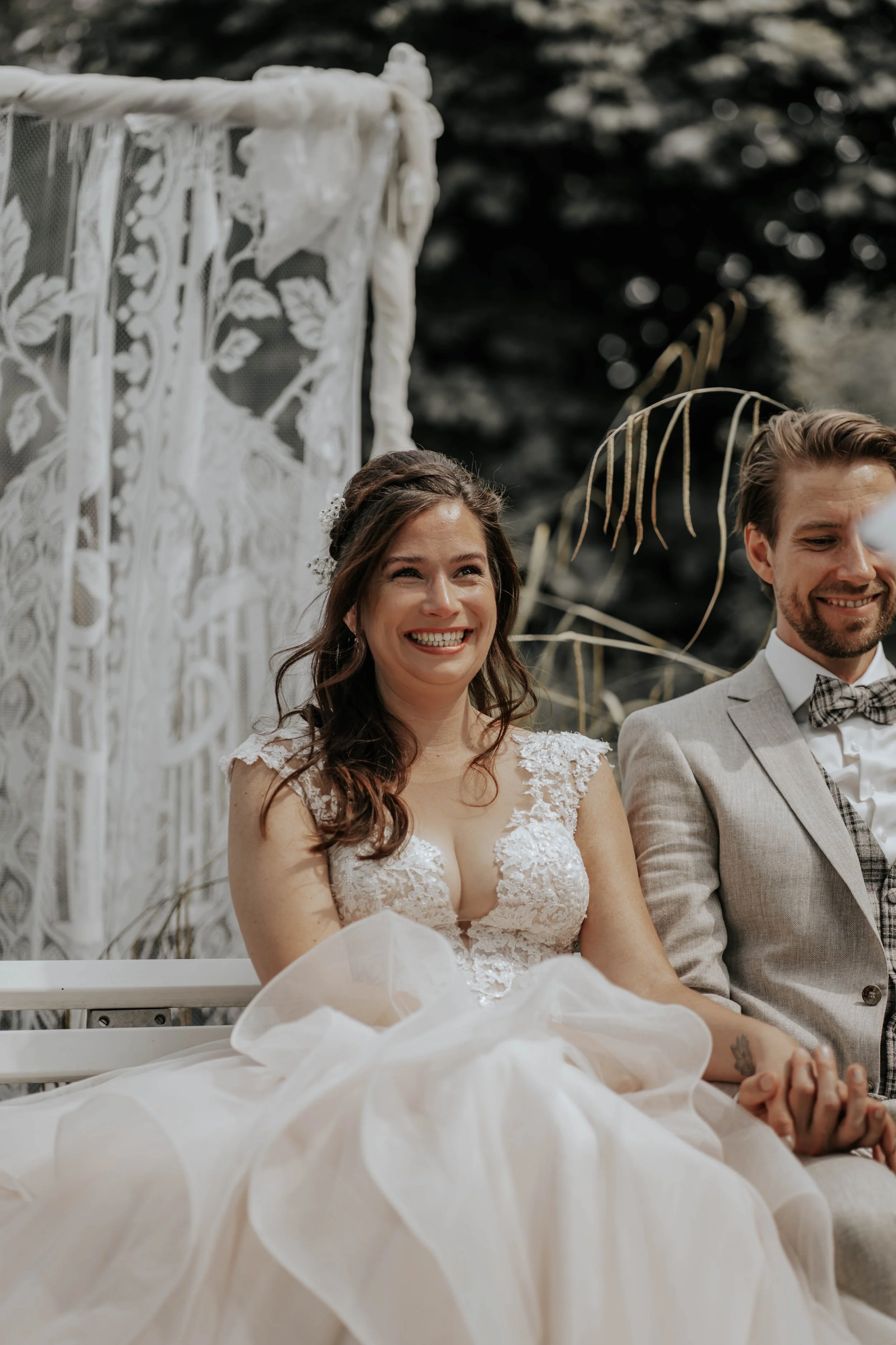 Een gelukkige bruid en bruidegom zitten samen buiten tijdens een huwelijksceremonie, met de bruid in een witte jurk met kanten details en de bruidegom in een lichtgrijs pak met een gestreepte strik. Op de achtergrond is decoratie met witte lace en na