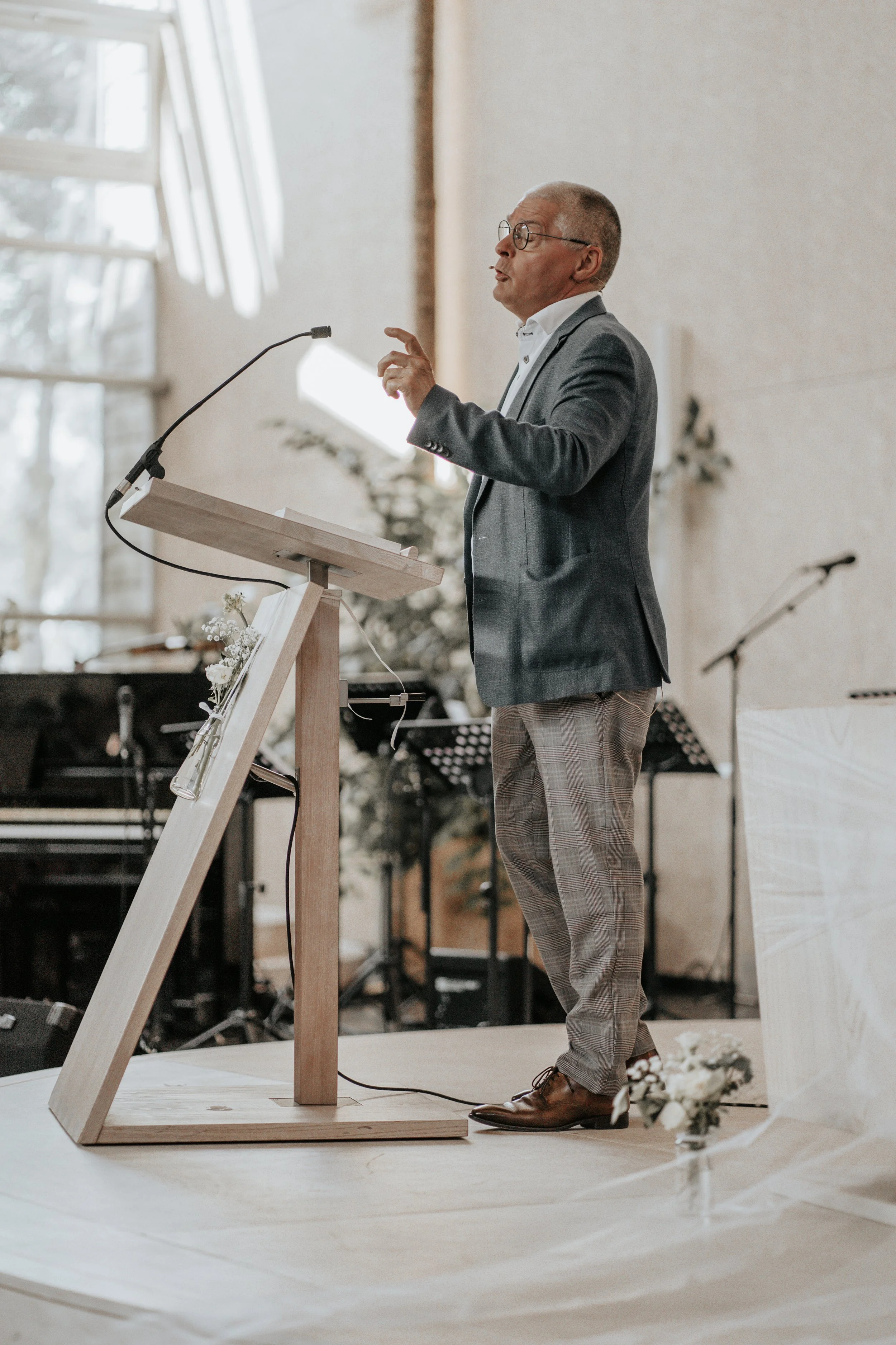 Man in een grijs pak geeft een presentatie achter een houten leestafel met microfoon in een lichte zaal.