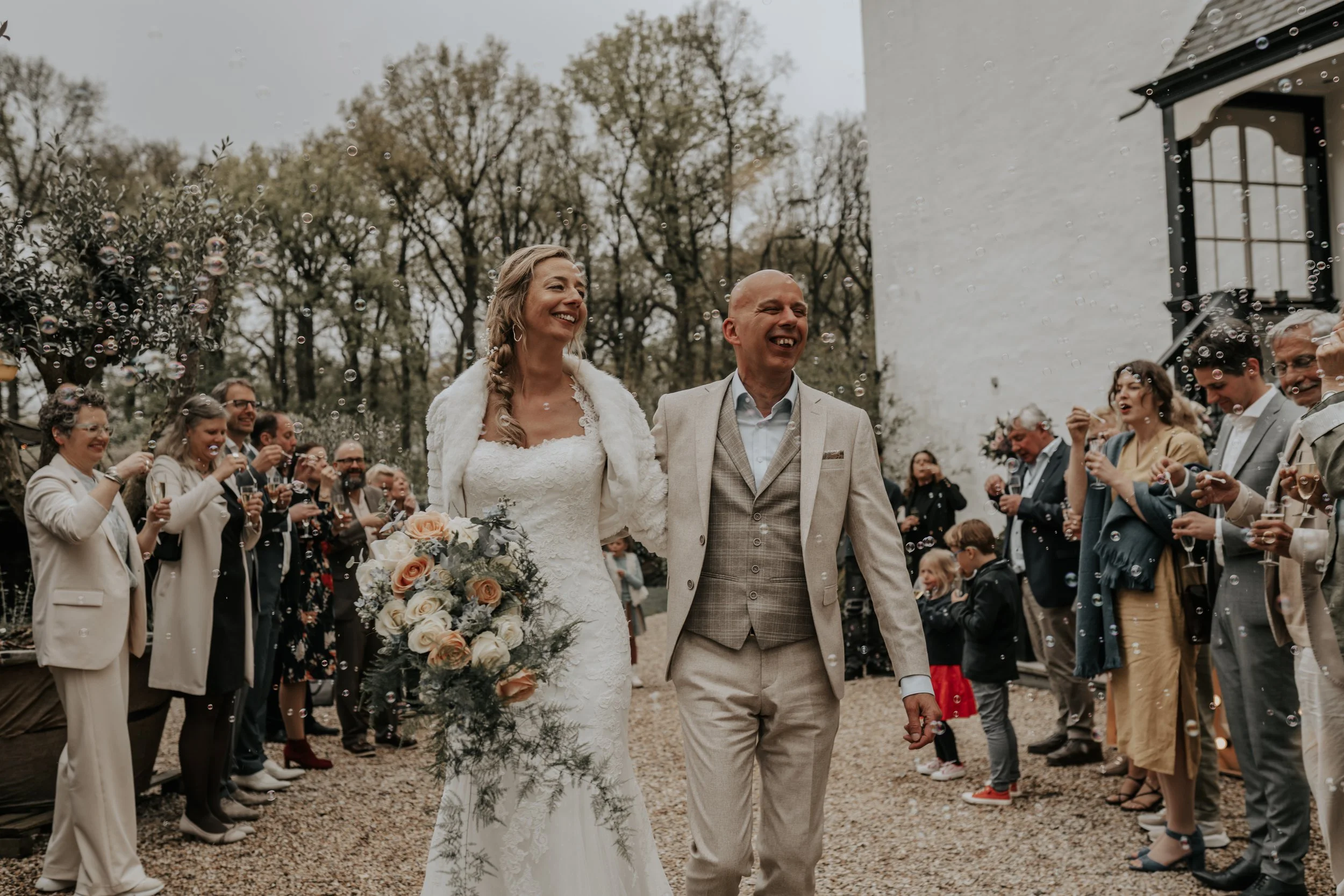 Bruid en bruidegom die samen lopen tijdens een huwelijksfeest, omringd door feestende gasten en belletjes, buiten onder een bewolkte hemel.