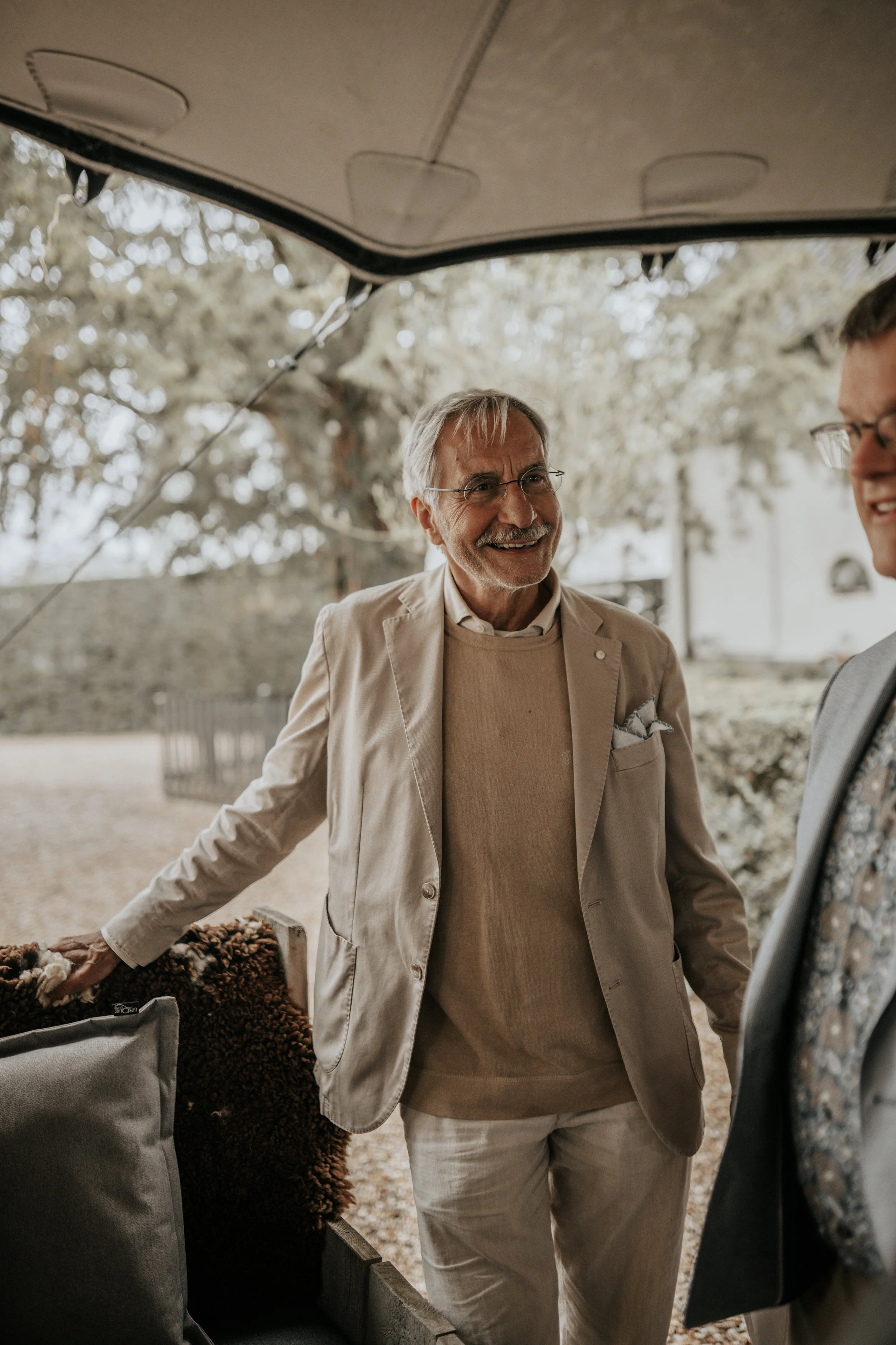 Twee mannen die in een open caravan of bus staan, lachend en met elkaar praten, buiten omgeven door bomen en een tuin, gekleed in lichte pakken.