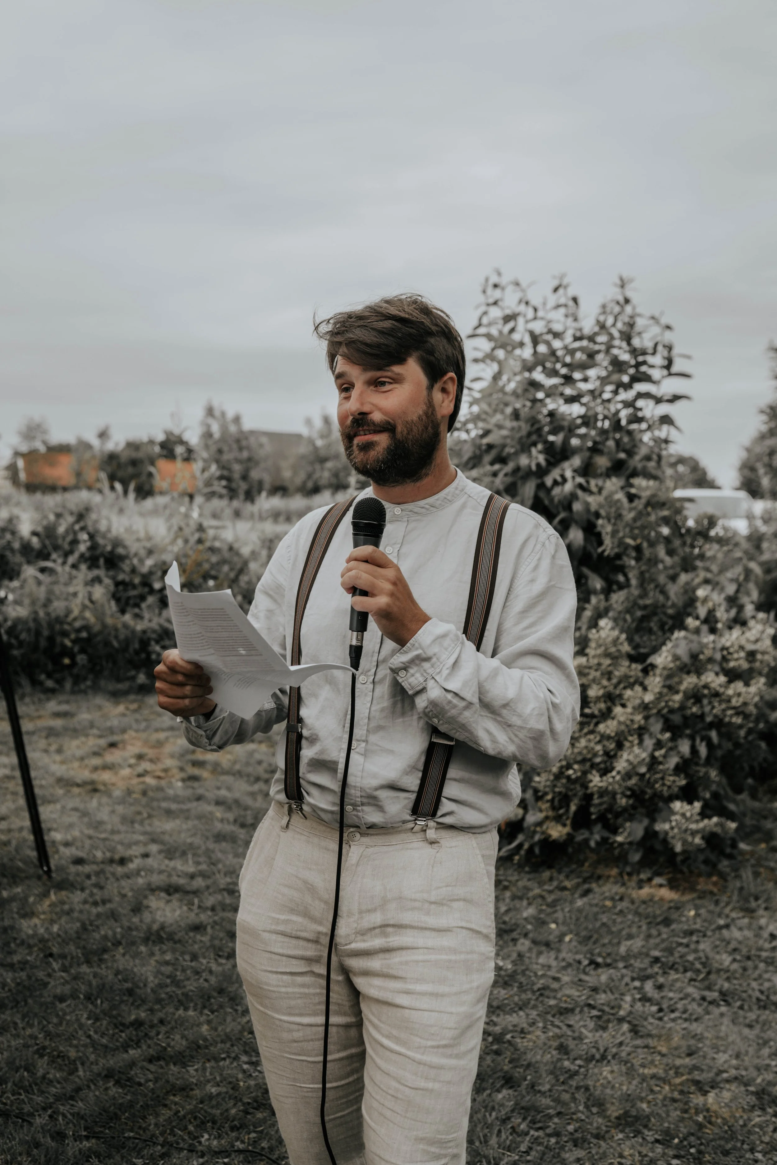Man houdt een speech in een tuin of park, hij heeft een microfoon en leest uit een stuk papier