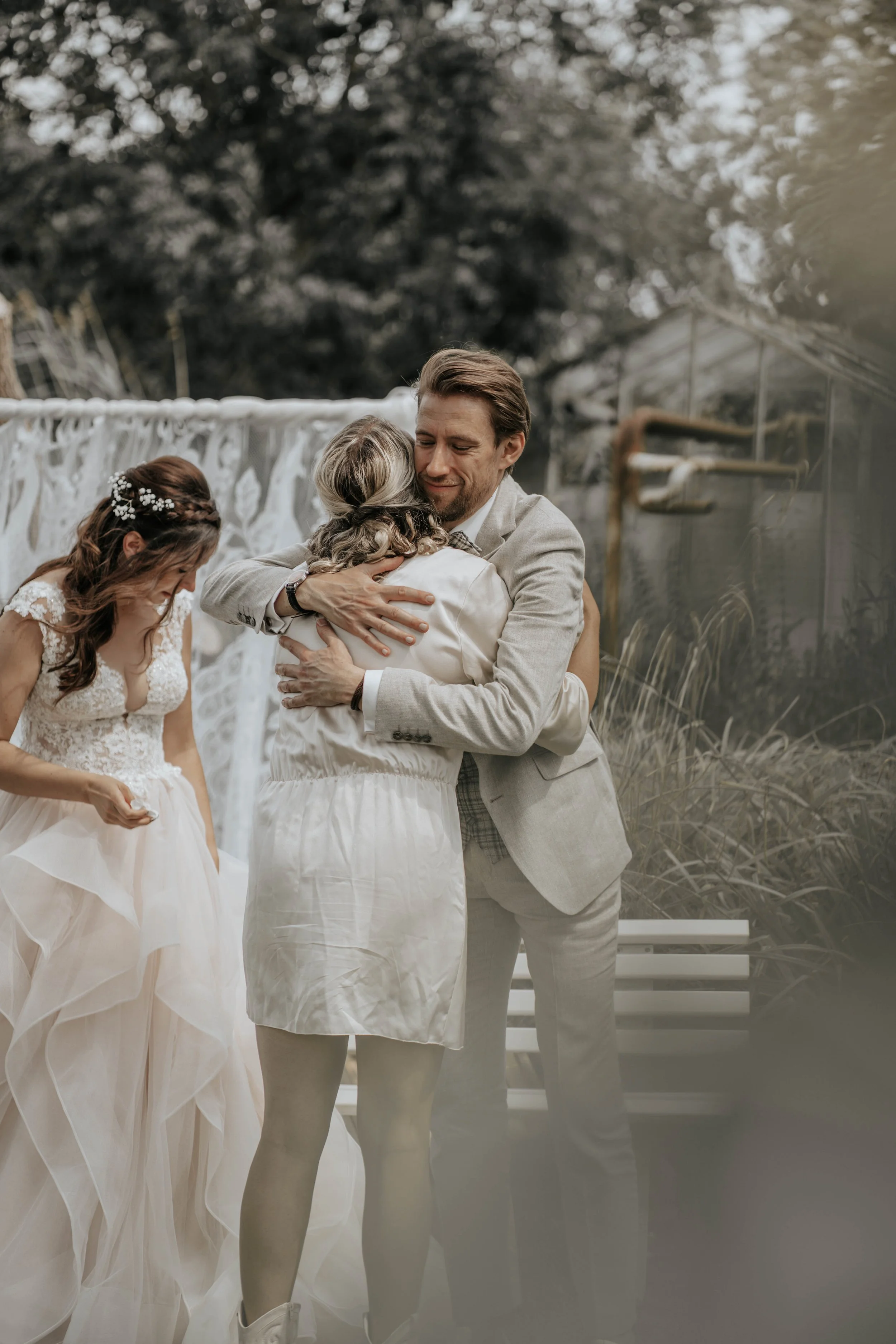 Een man en een vrouw omhelzen elkaar tijdens een bruiloft, terwijl een andere vrouw in een trouwjurk staat te lachen en te kijken.
