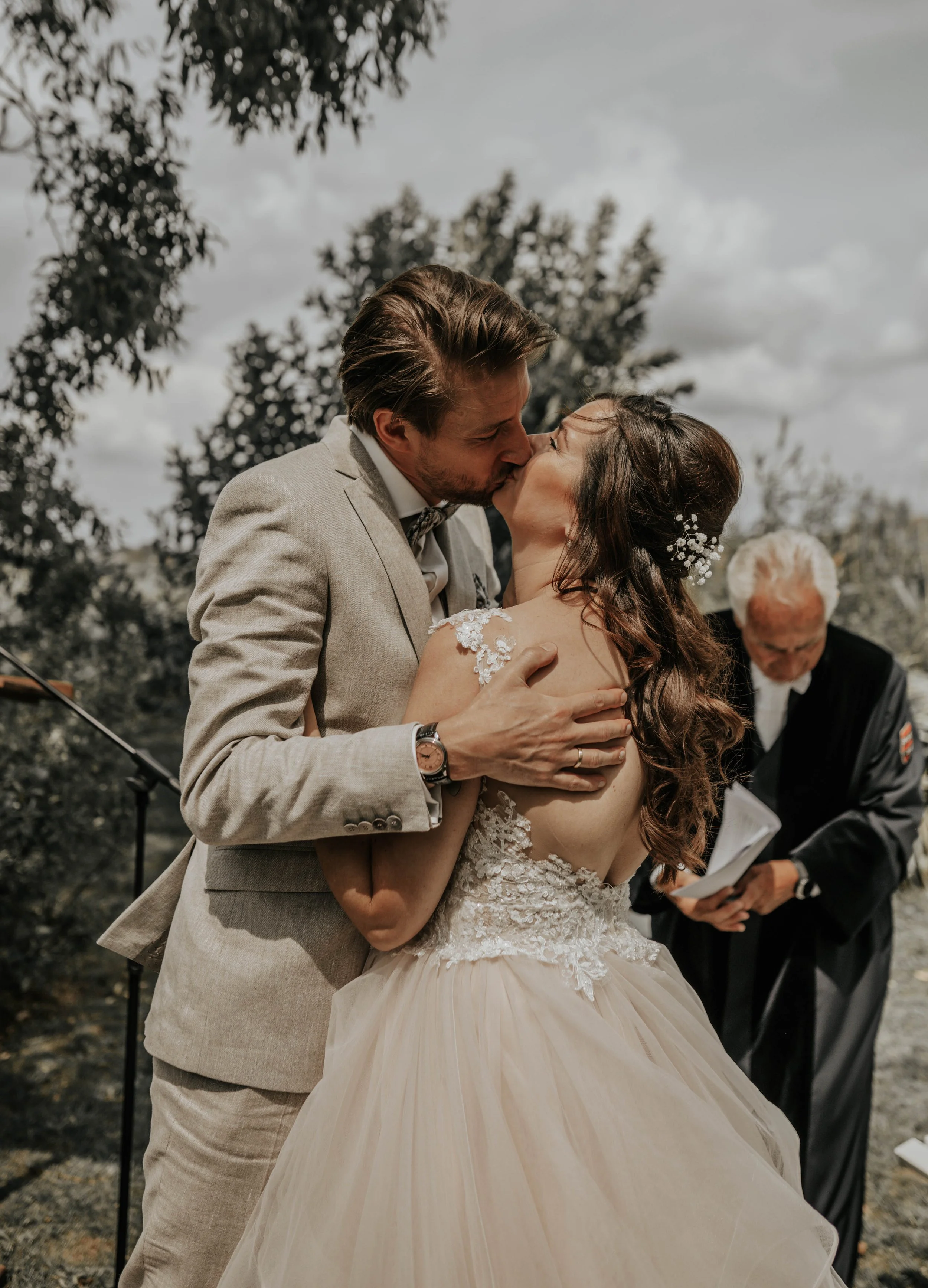 Een bruidskoppel kust elkaar tijdens hun huwelijk, met een ceremoniemeester op de achtergrond, outdoors, met bomen en een bewolkte hemel.