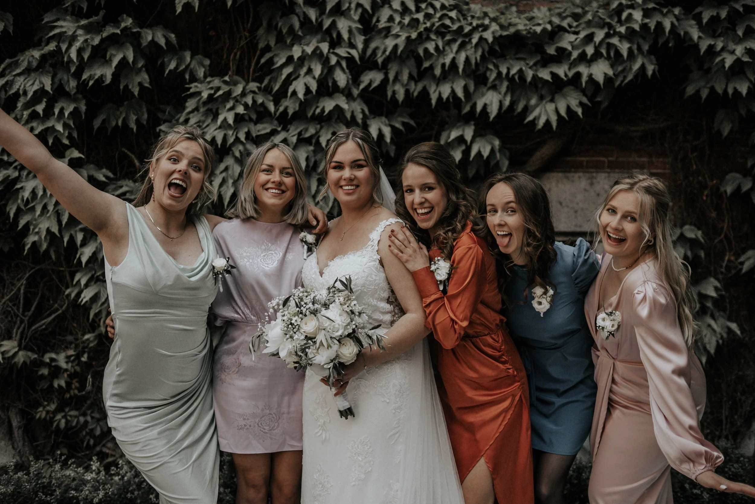 Vrouw in witte trouwjurk met bloemboeket, omringd door zes vrouwen in verschillende gekleurde jurken, vriendinnen op een bruiloft.