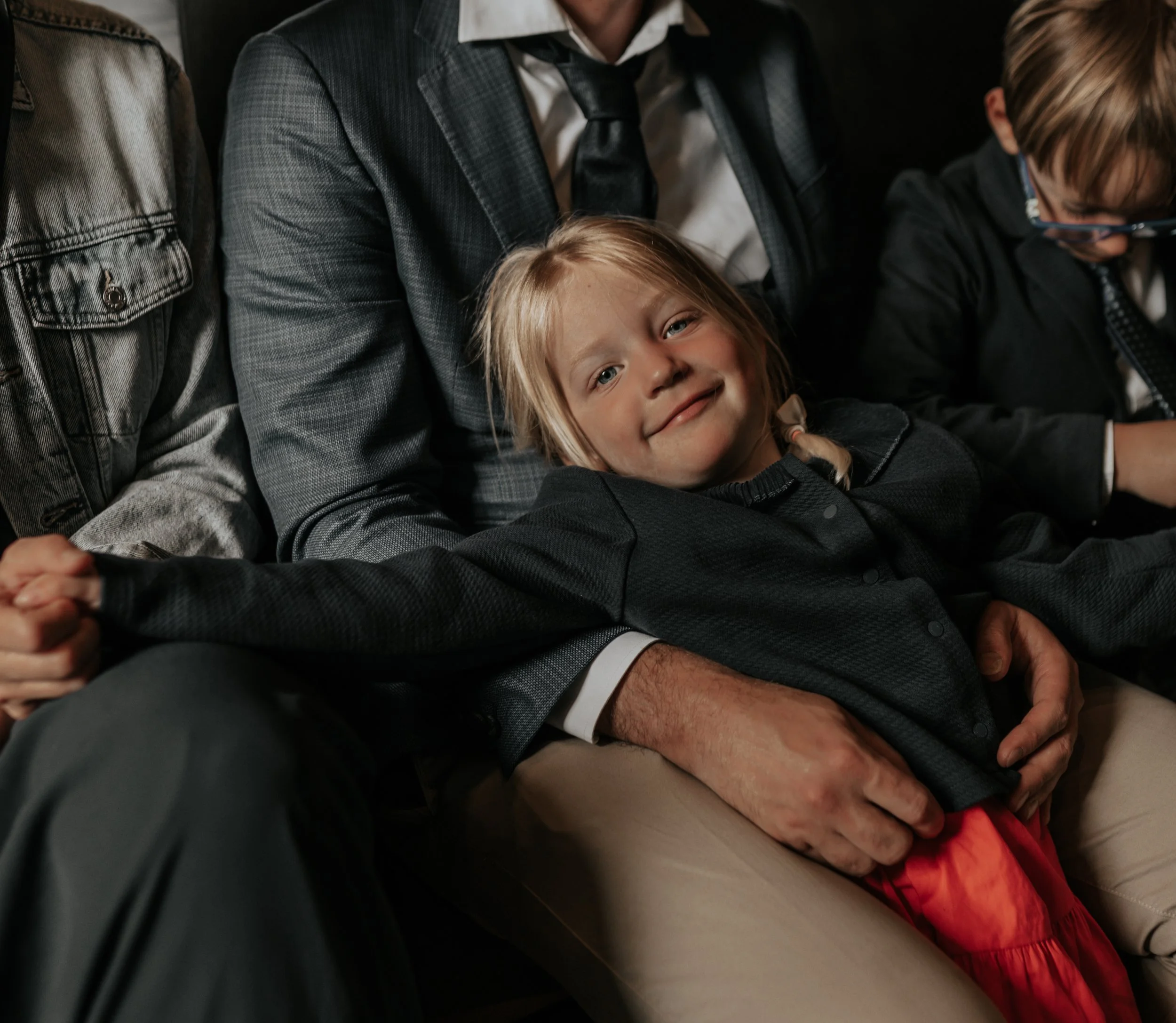 Een jonge meisje glimlacht terwijl ze op schoot zit van een man in een grijs pak, omringd door andere kinderen en volwassenen.