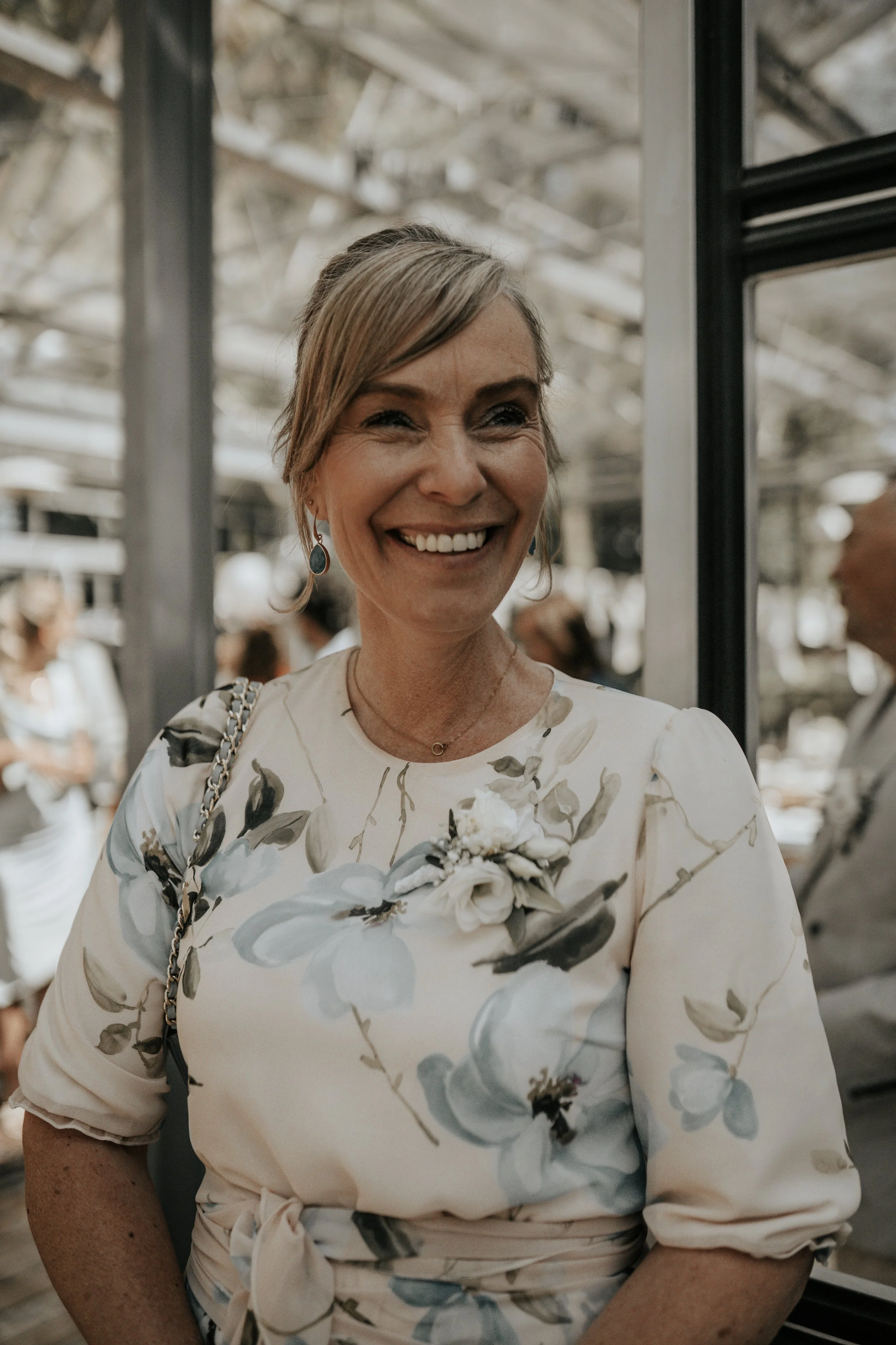 Een lachende vrouw met blond haar in een bloemenjurk met een bloemcorsage, mogelijk op een feest of bruiloft.