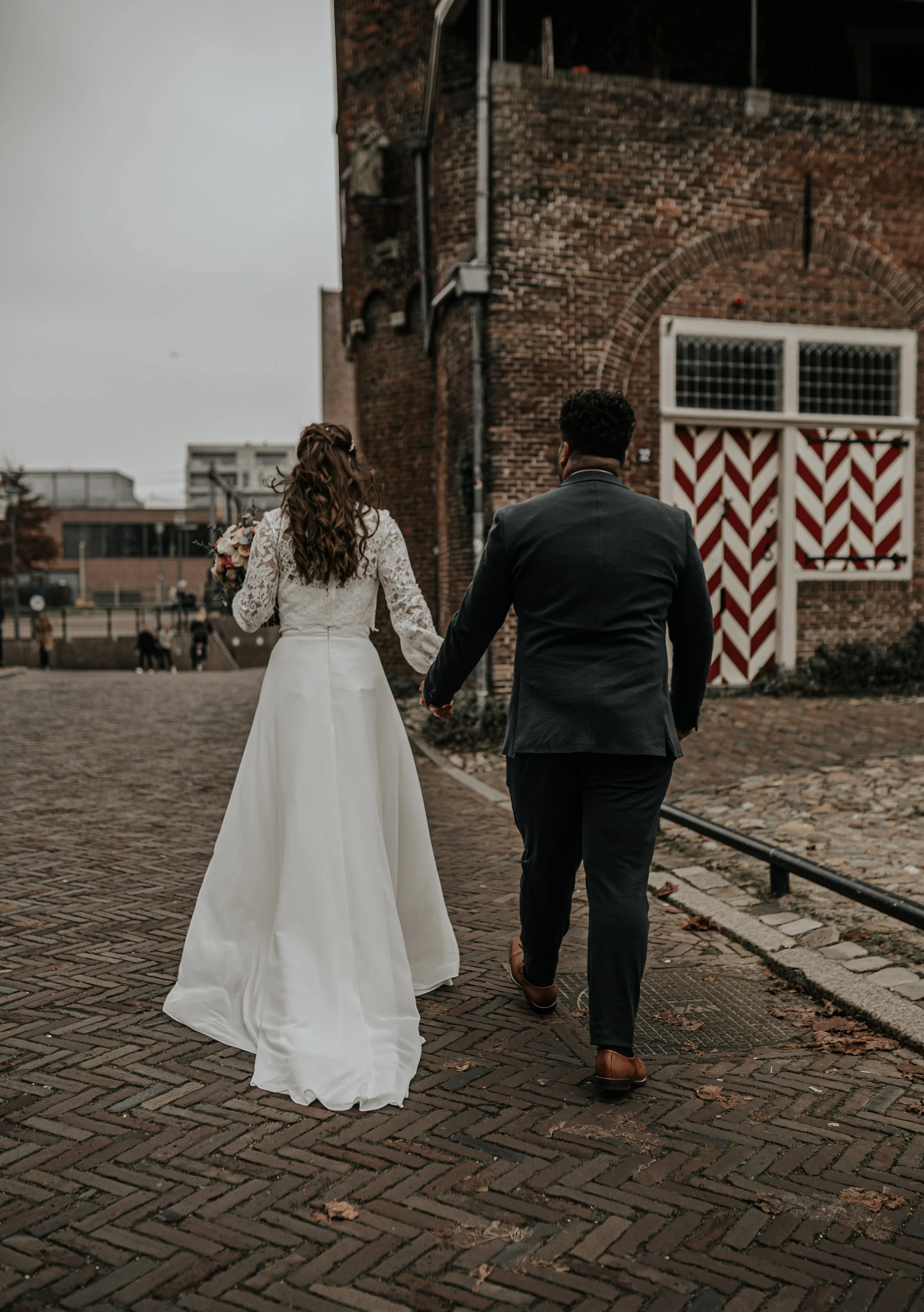 Een getrouwde paar loopt hand in hand door een straat, met een man in een zwart pak en een vrouw in een witte trouwjurk, onder een bewolkte hemel.