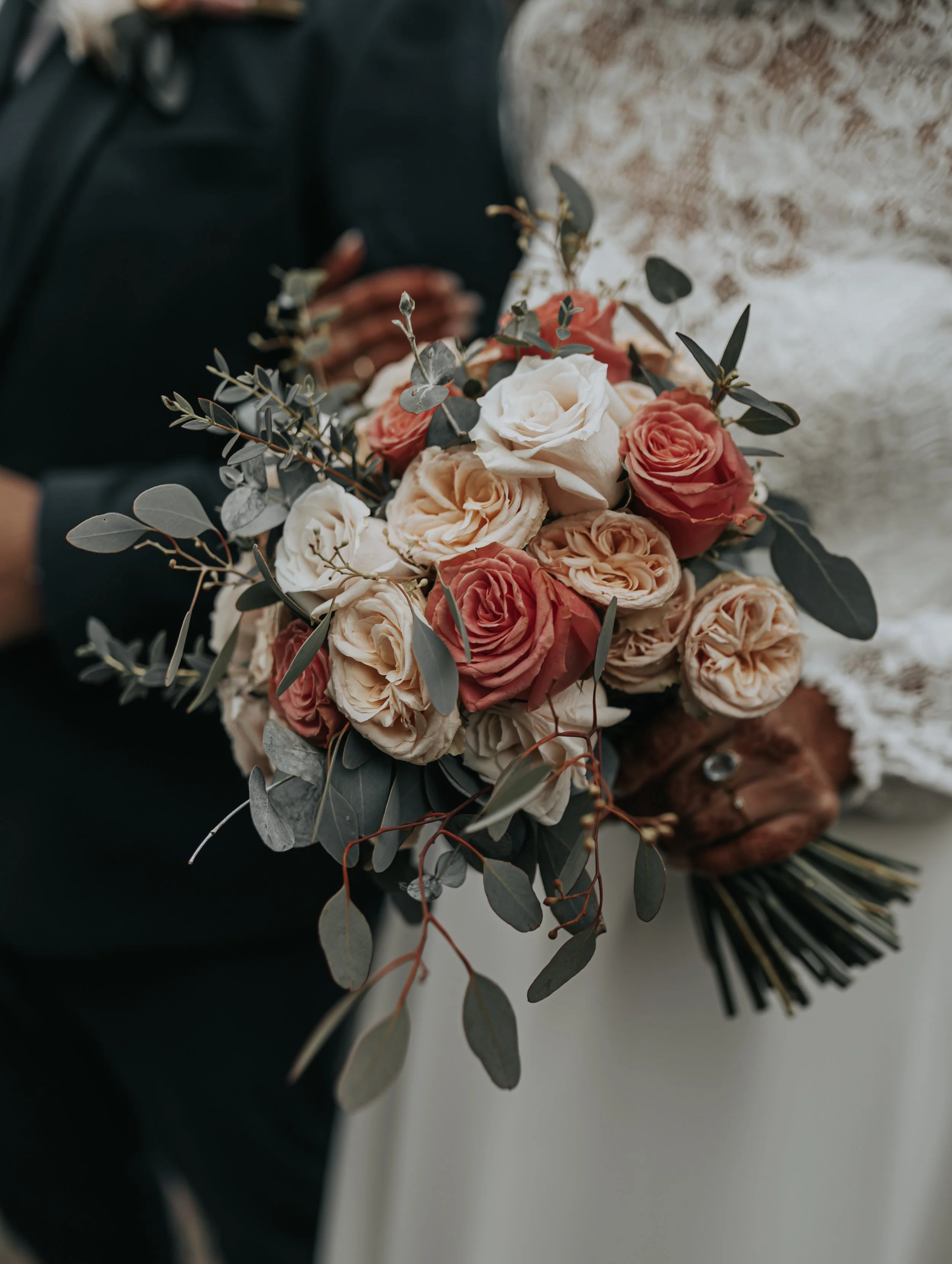 Close-up van een bruid met een bloemenboeket bestaande uit witte en roze rozen en groen blad, vastgehouden door een persoon in een zwarte outfit.