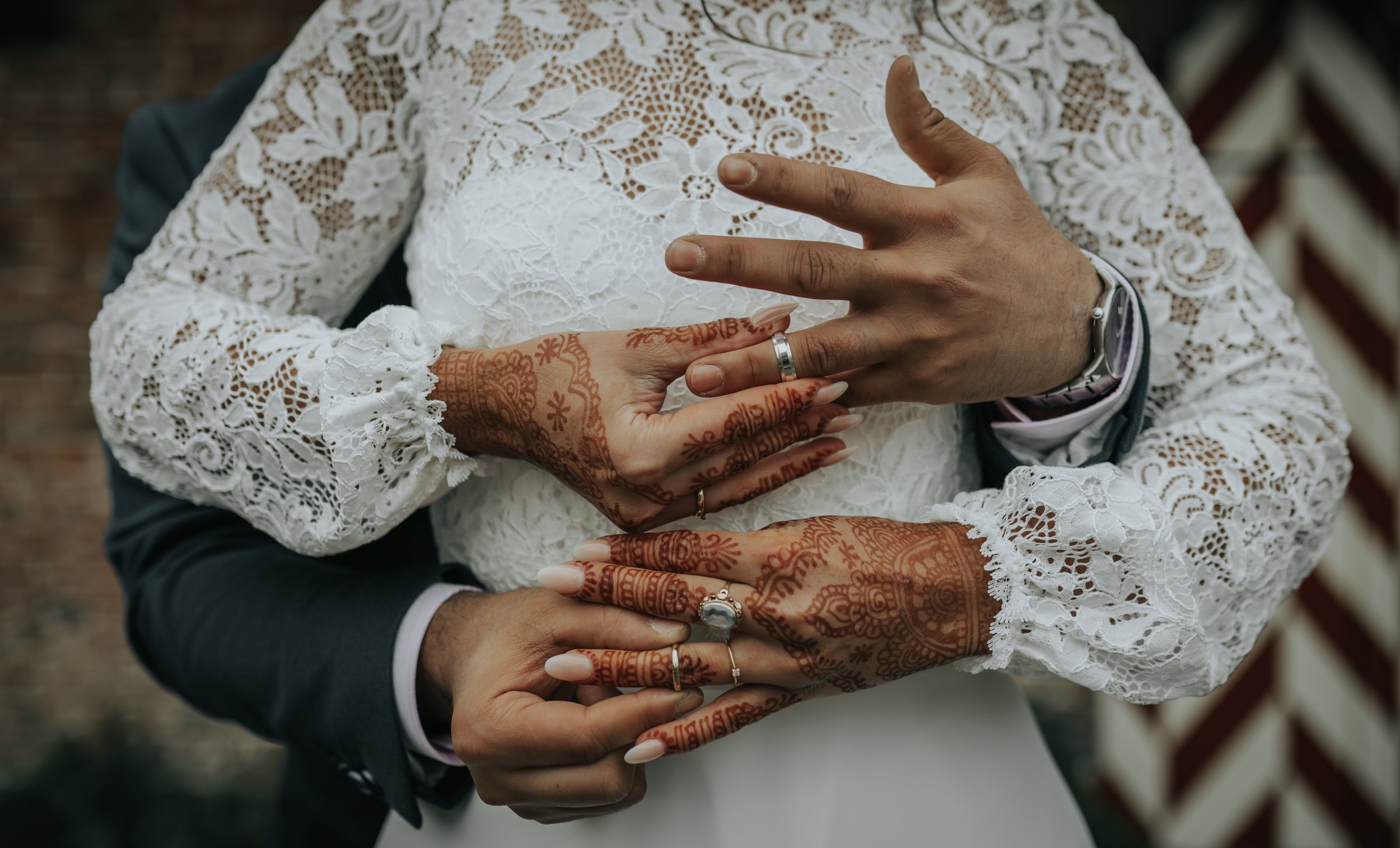 Close-up van een bruid en bruidegom die elkaar vasthouden, waarbij de handen van de bruid met henna en sieraden zichtbaar zijn, en de bruid een witte kanten jurk draagt.