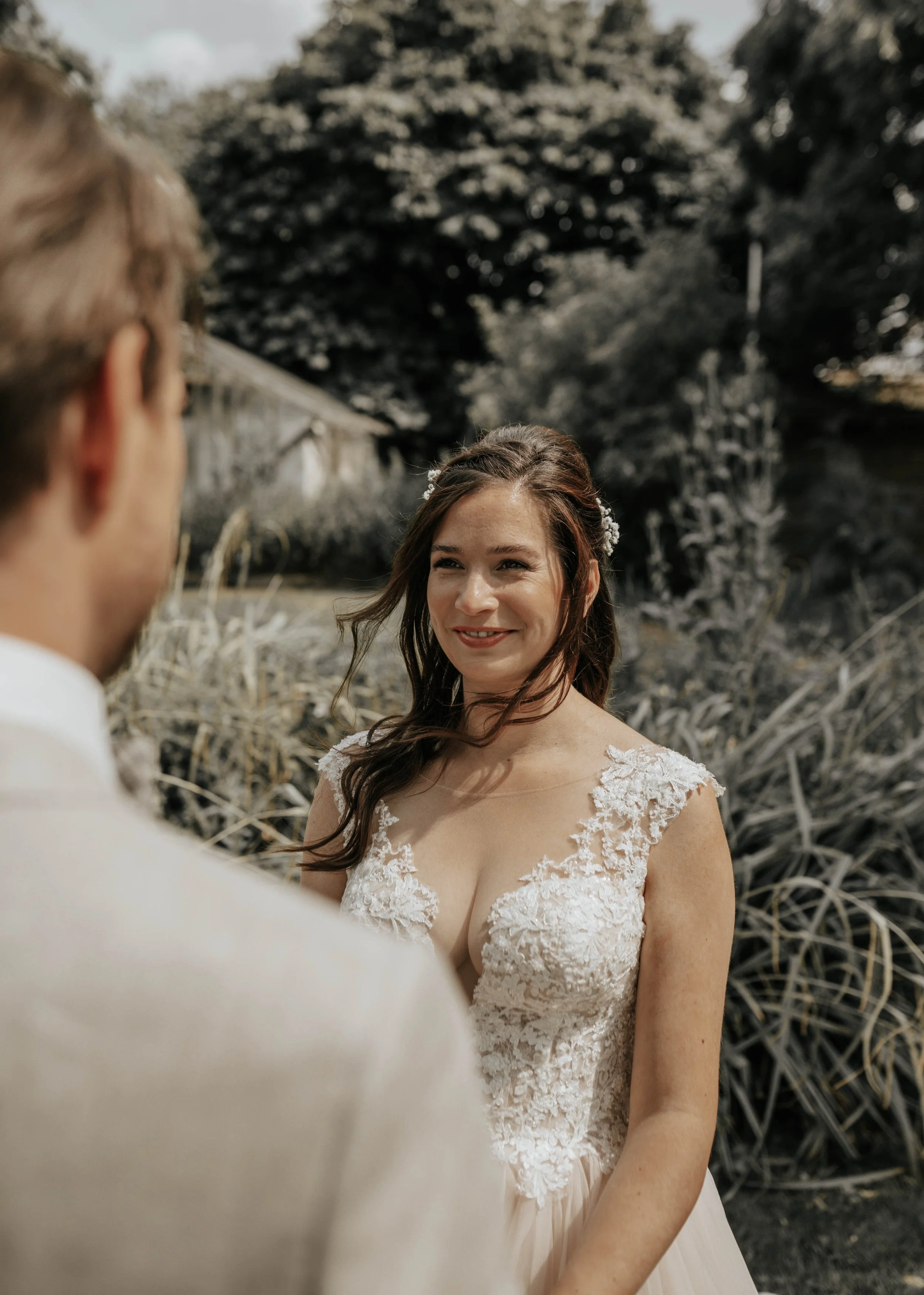 Een bruiloftsfoto van een vrouw in een witte kanten trouwjurk die naar een man kijkt, buiten in een natuurlijke omgeving met bomen en struiken.