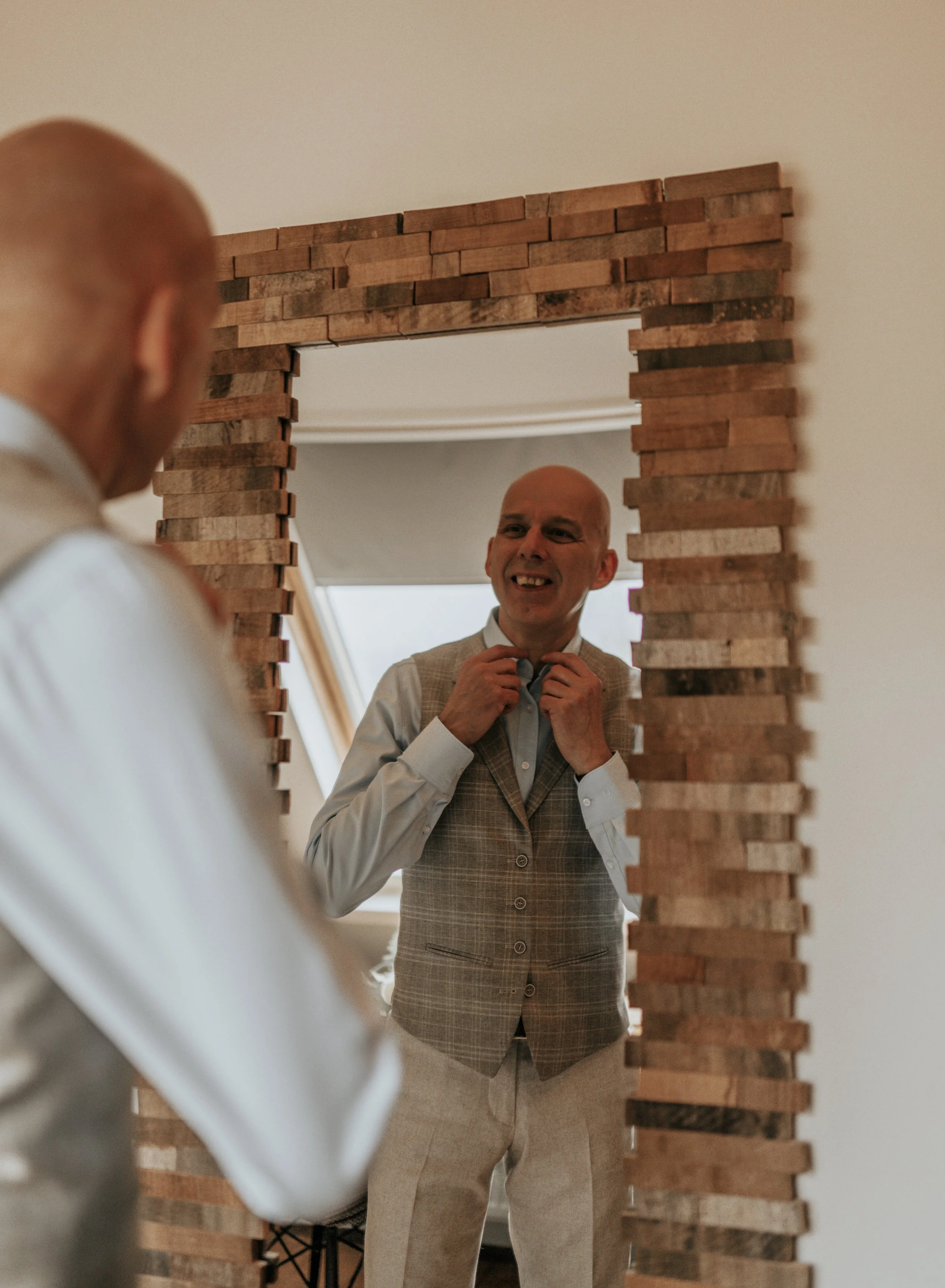 Man die zichzelf in een houten ombouwde spiegel bekijkt, gekleed in formele kleding, en zichzelf helpt met de knoop van een hemd.