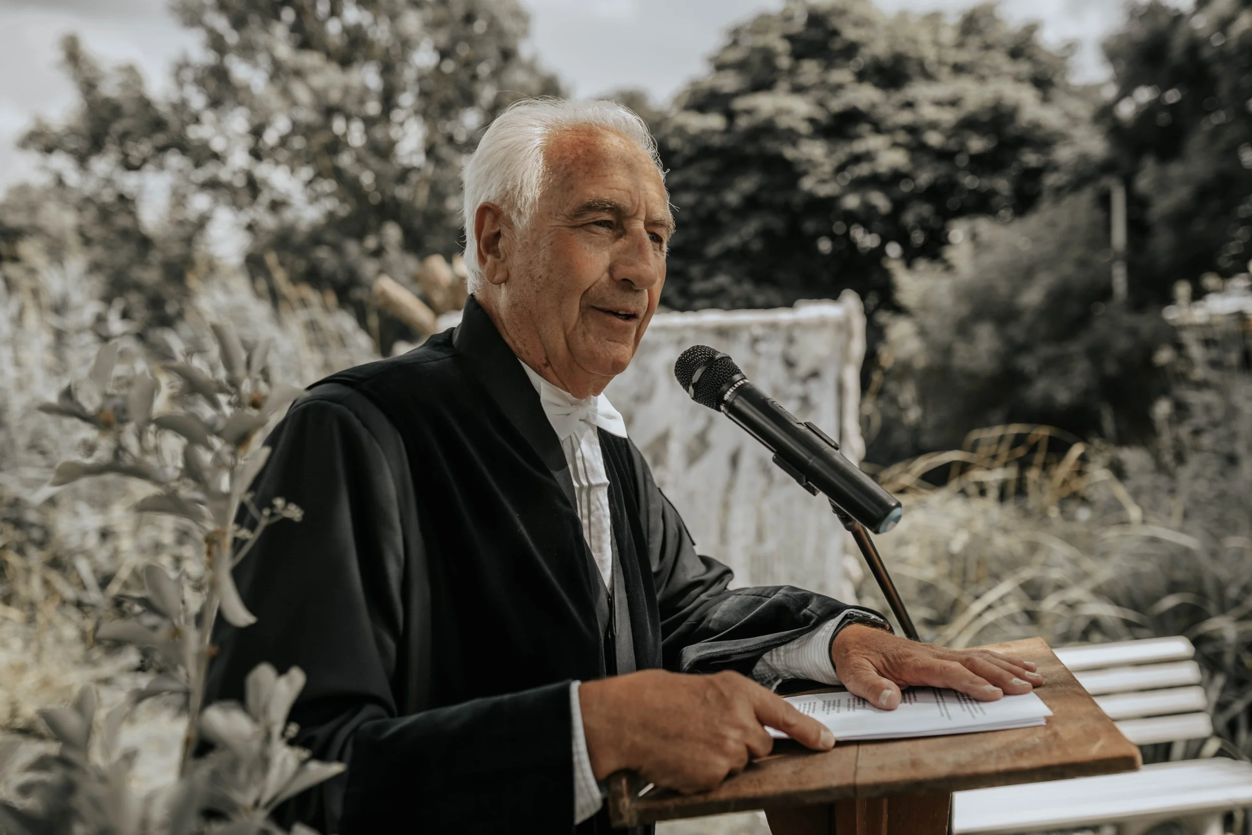 Een oudere man in een toga leest voor vanaf een houten verhoogje tijdens een buitenceremonie, met een microfoon voor hem en planten op de voorgrond.