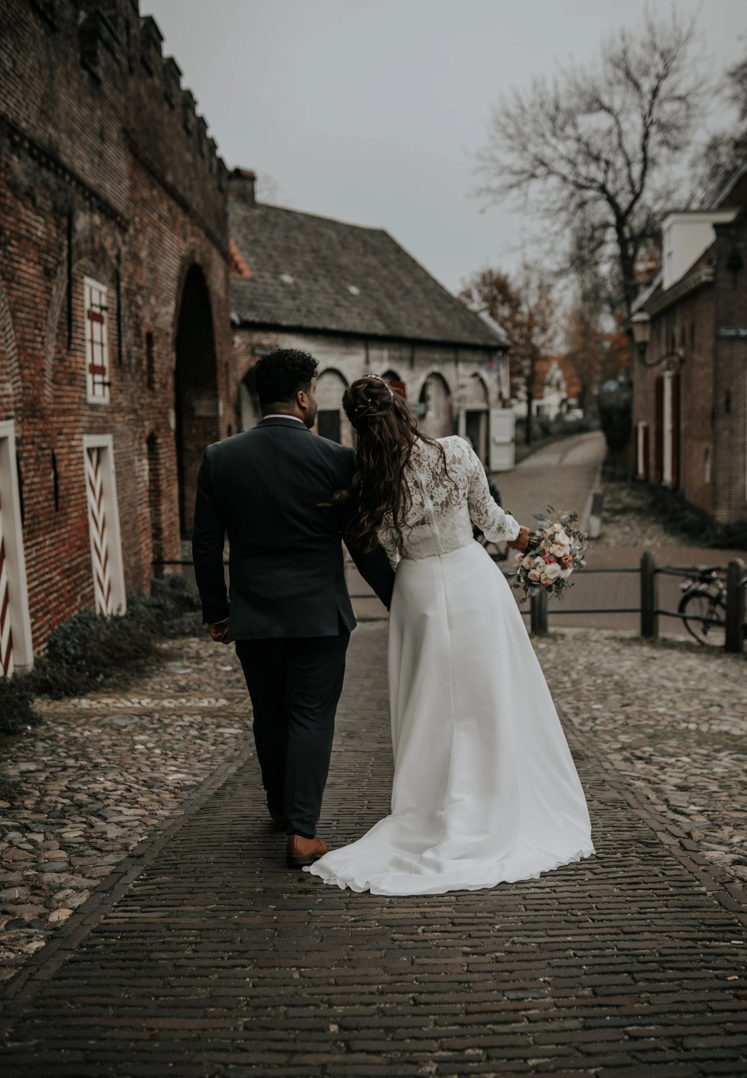 Een koppel in trouwkleding loopt hand in hand door een oude straat met stenen loop en historische gebouwen, mogelijk voor een bruiloft.
