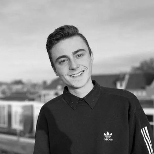 Een jonge man met bril en een zwarte Adidas-trui staat buiten, lachend met gebouwen en een trein op de achtergrond.