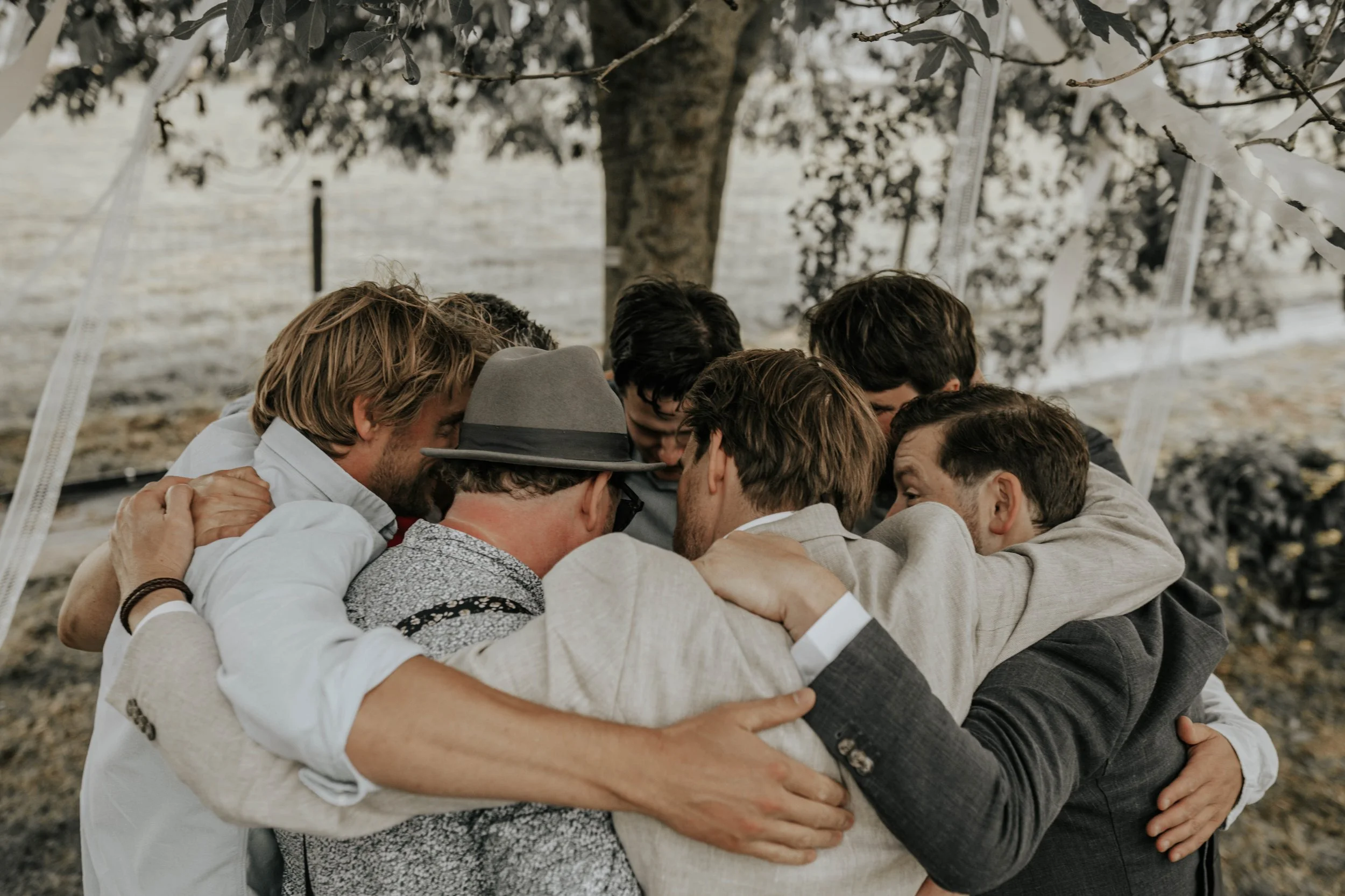 Een groep mannen die in een cirkel staan en elkaar omarmen, onder een boom.