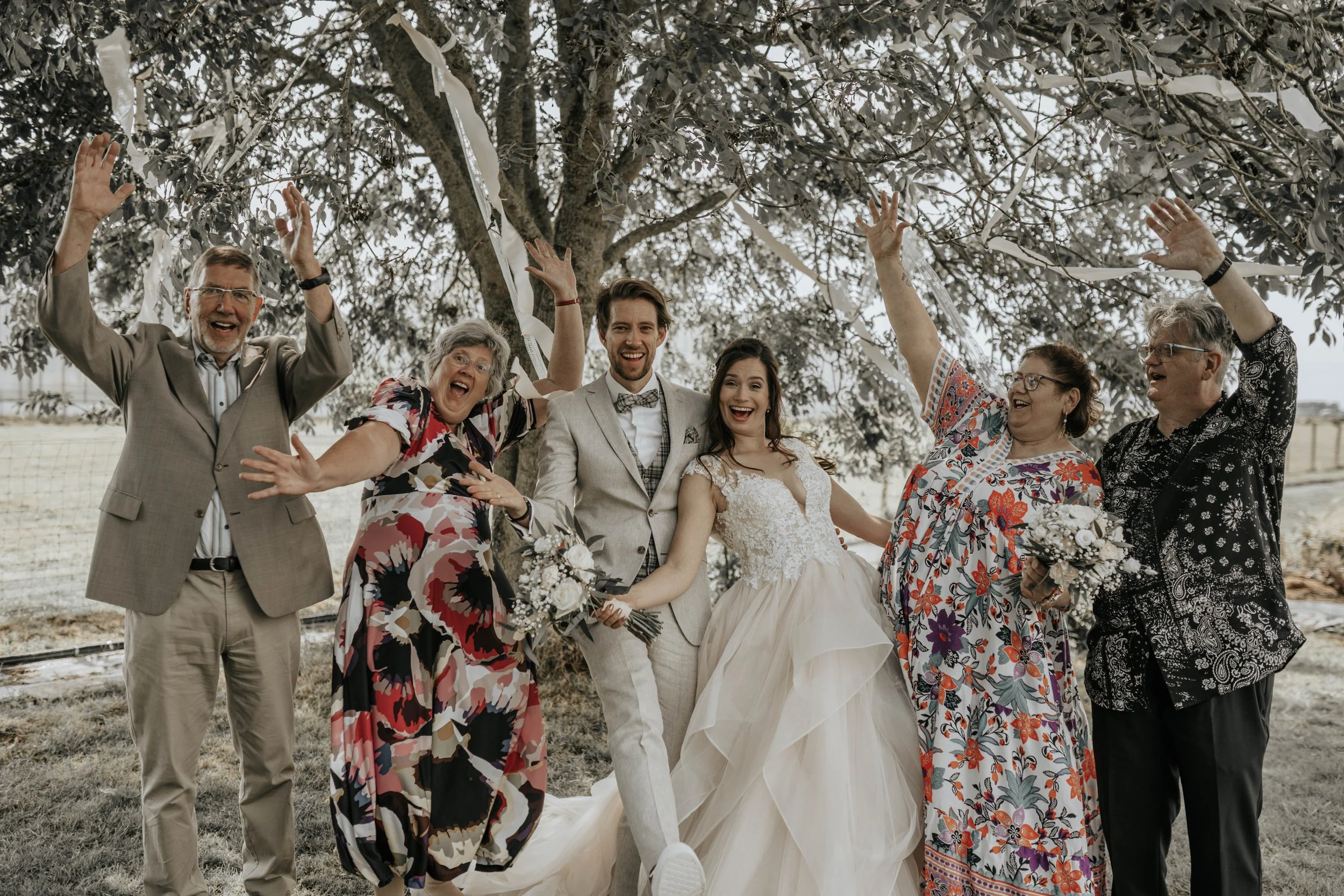 Bruid en bruidegom samen met vier oudere familieleden onder een boom, lachend en feestend, in een buitengebeuren.